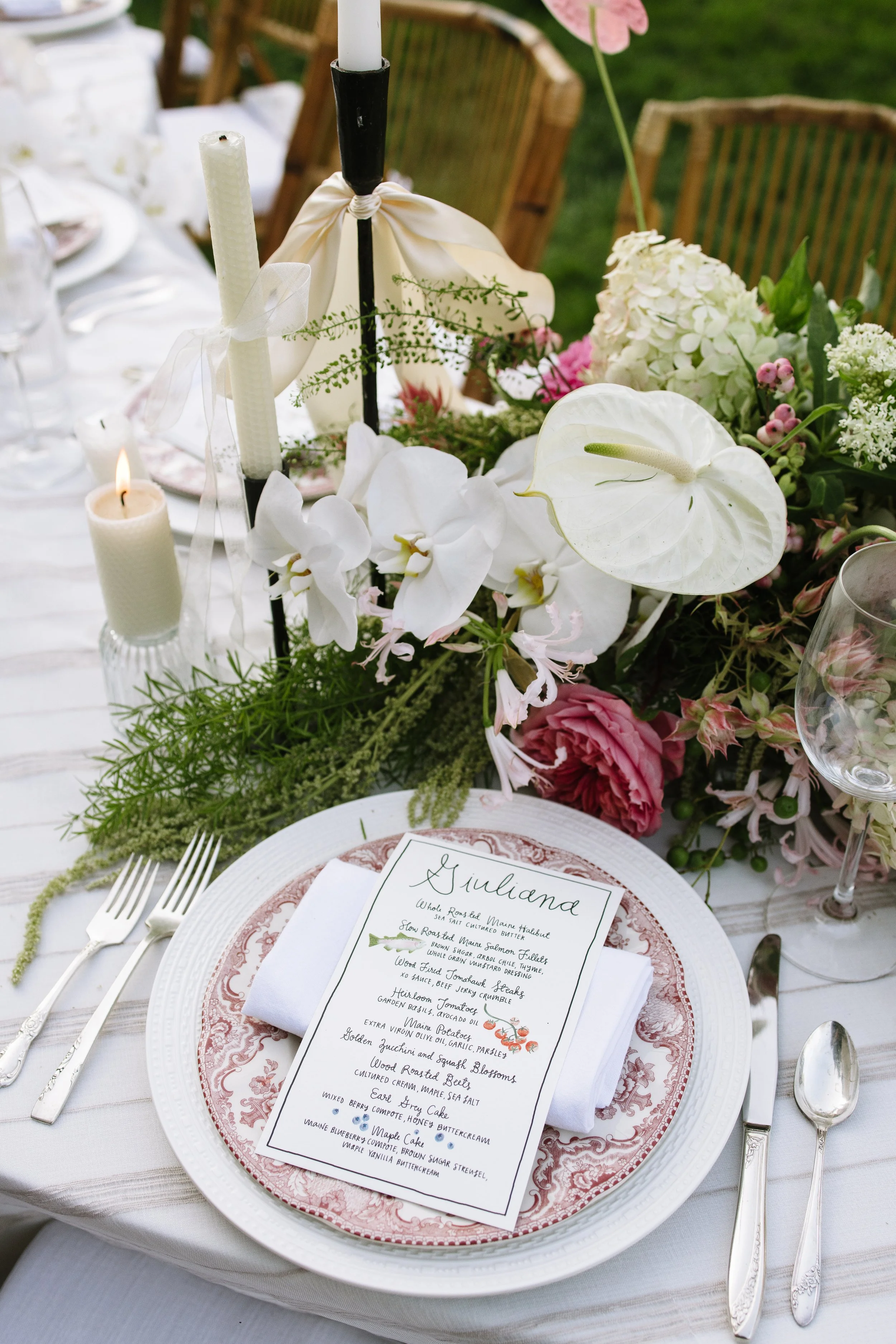 Elegant outdoor table setting with a floral centerpiece featuring white orchids, anthuriums, and pink flowers. A white plate with a floral-patterned charger and a handwritten menu card is in the foreground. Silverware is arranged neatly, and candles 