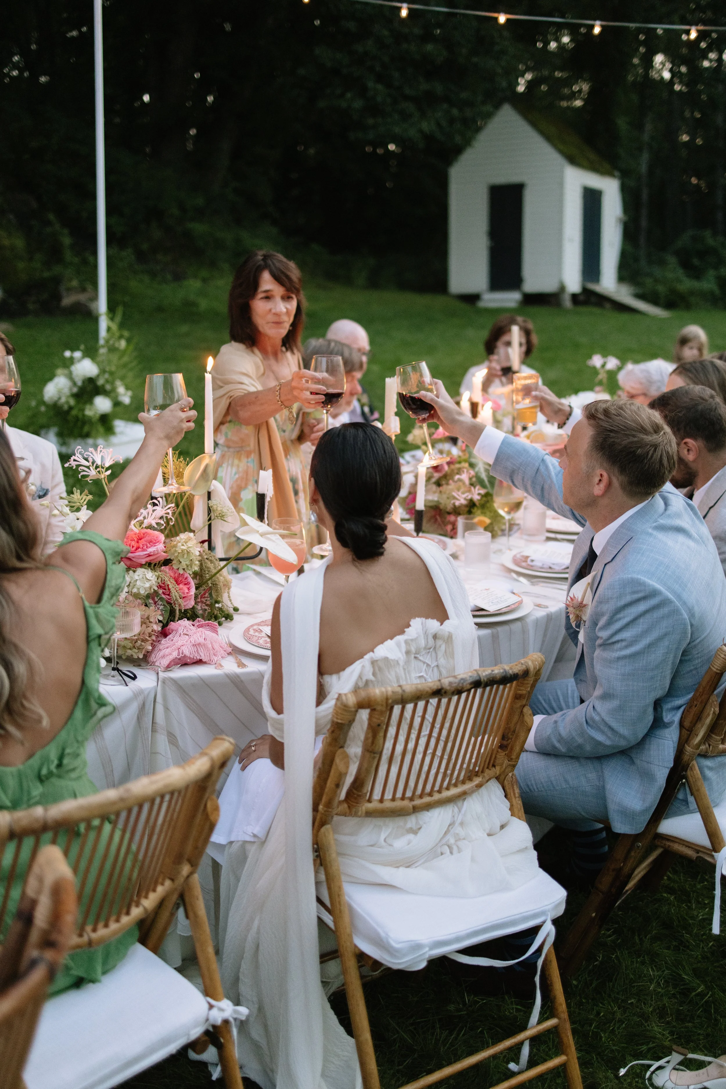 People gathered around a dinner table outdoors raising glasses in a toast during sunset at a celebration, with a small white shed in the background.