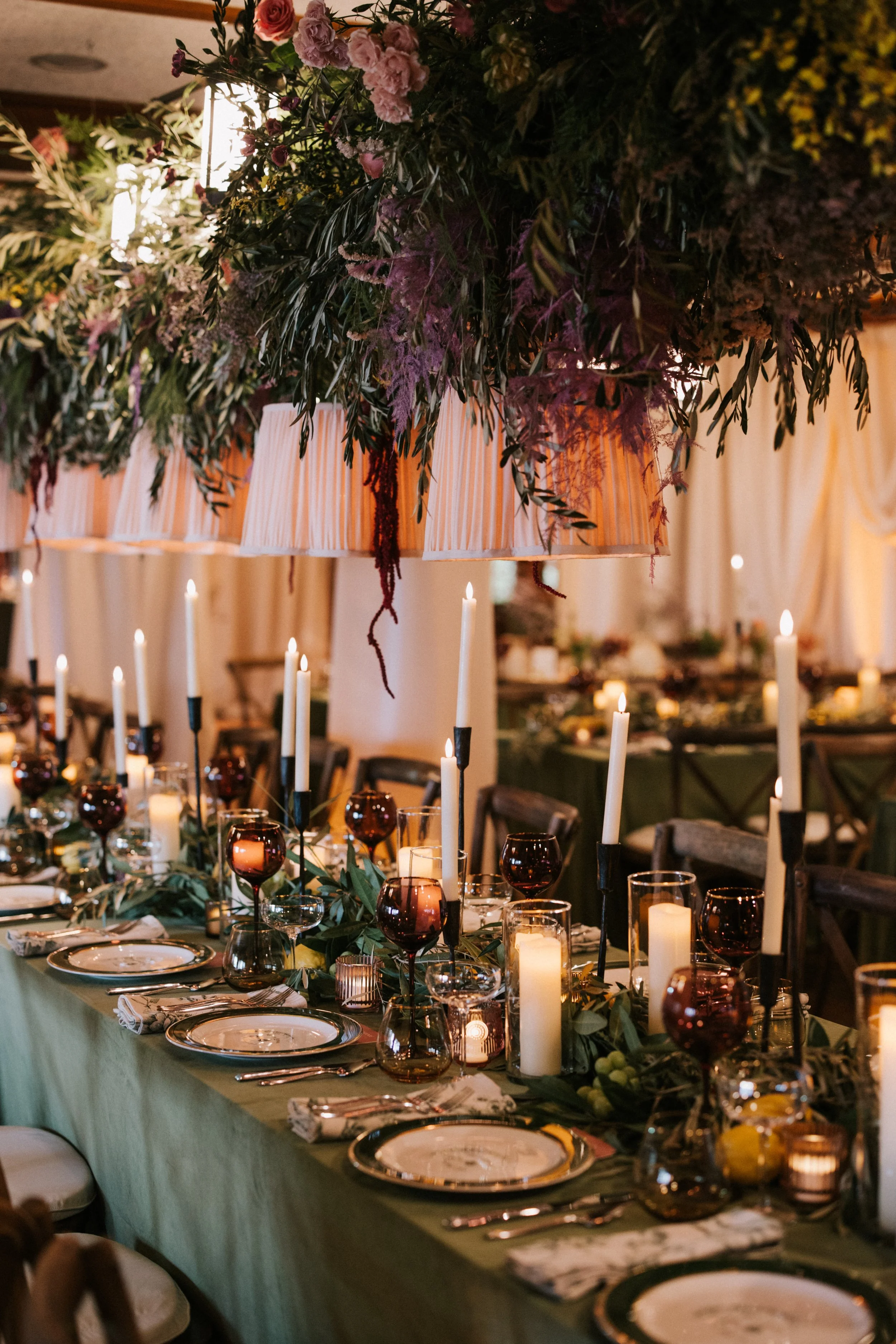 Elegant banquet table decorated with candles, wine glasses, and greenery, with a large floral centerpiece hanging overhead.