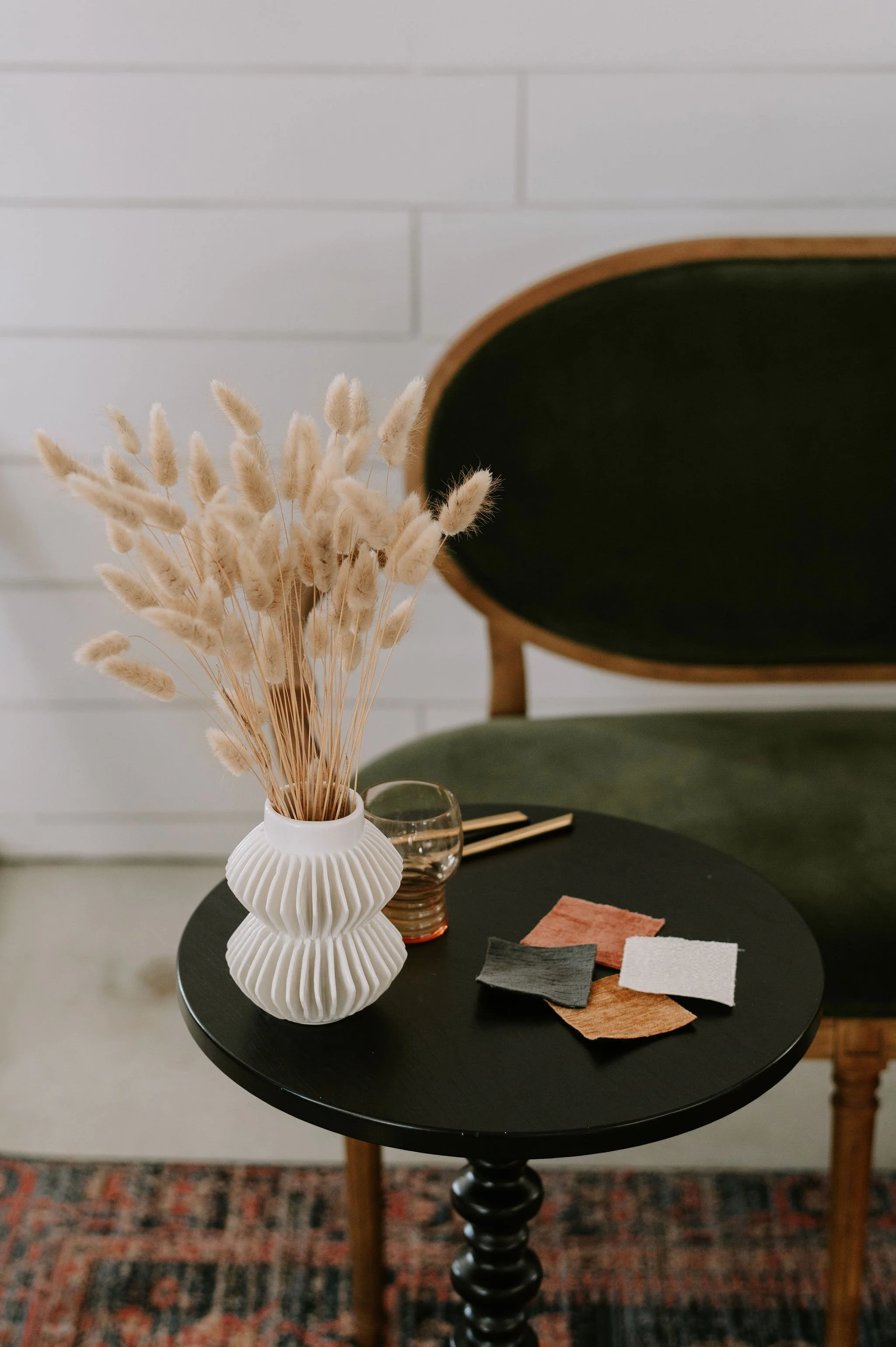 A black round table with a white ceramic vase containing dried beige grass, a small glass with a red tint, a pair of metallic chopsticks, and fabric swatches in black, pink, white, and brown. Behind the table is a green velvet sofa with a wooden frame, and a white brick wall in the background.