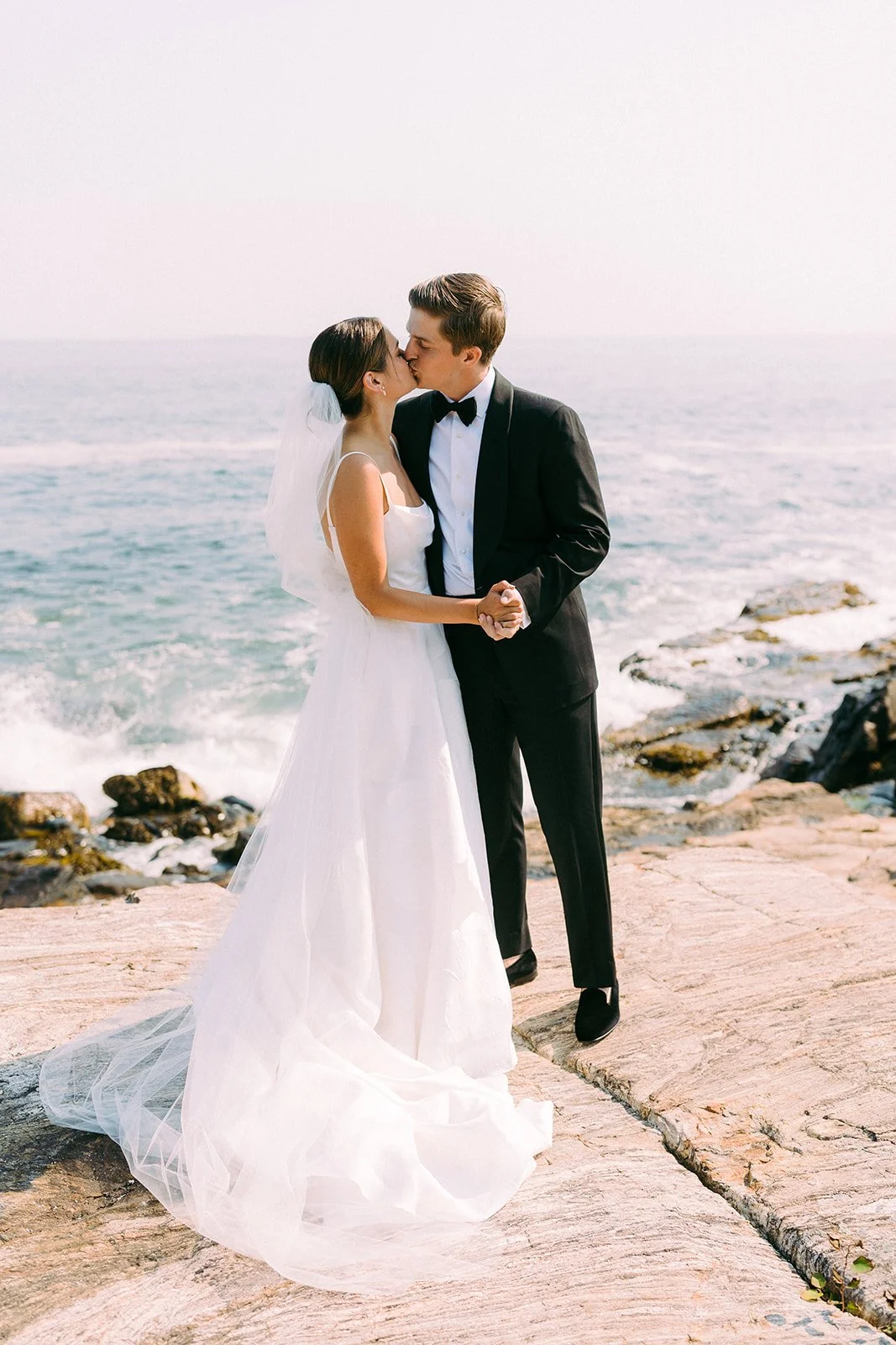 A bride and groom share a kiss on a rocky beach, with the ocean in the background, during their wedding.