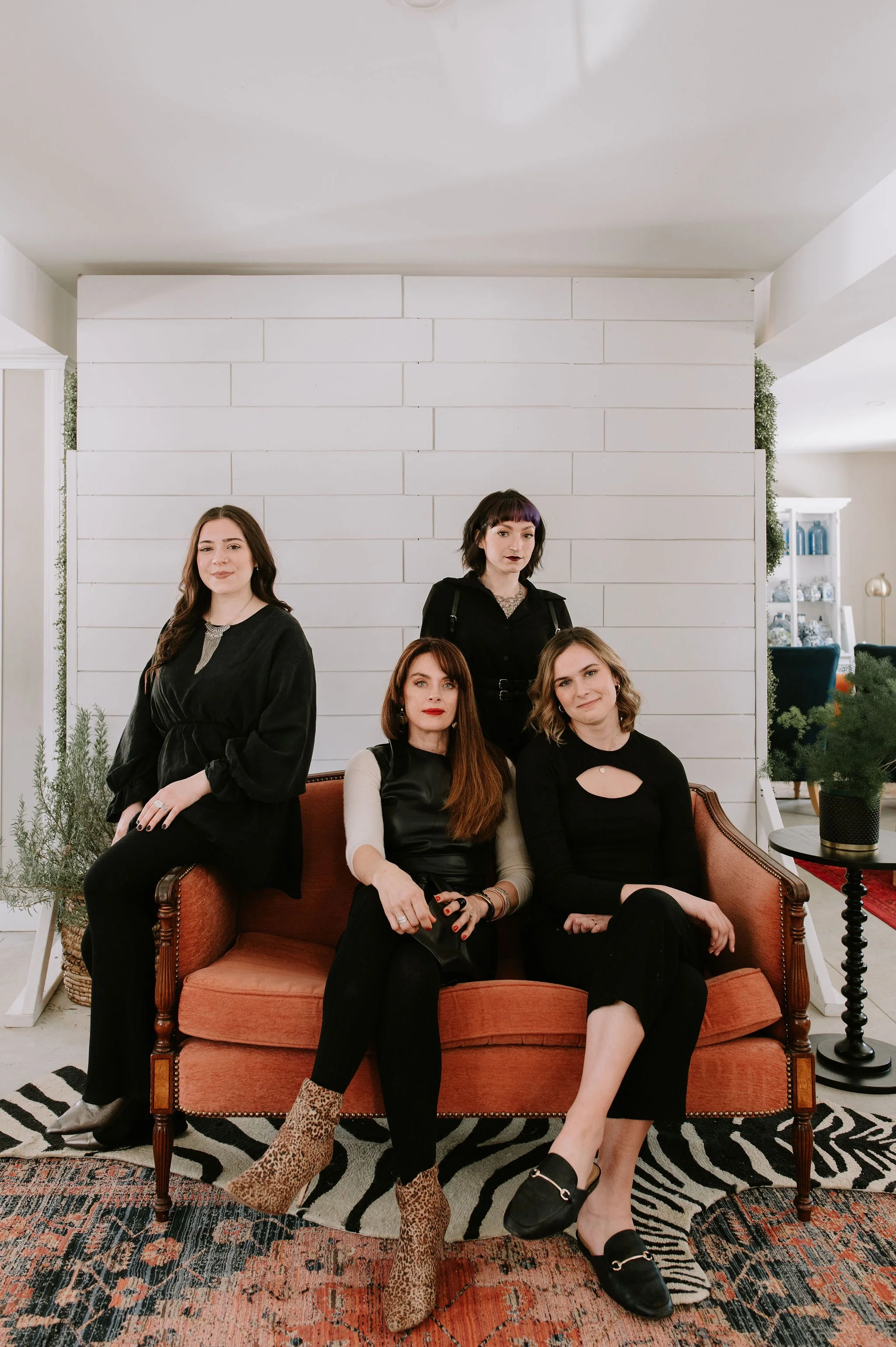 Four women with varying hair colors and styles posing in a living room with white shiplap wall, plants, and furniture, dressed in black and neutral colors.