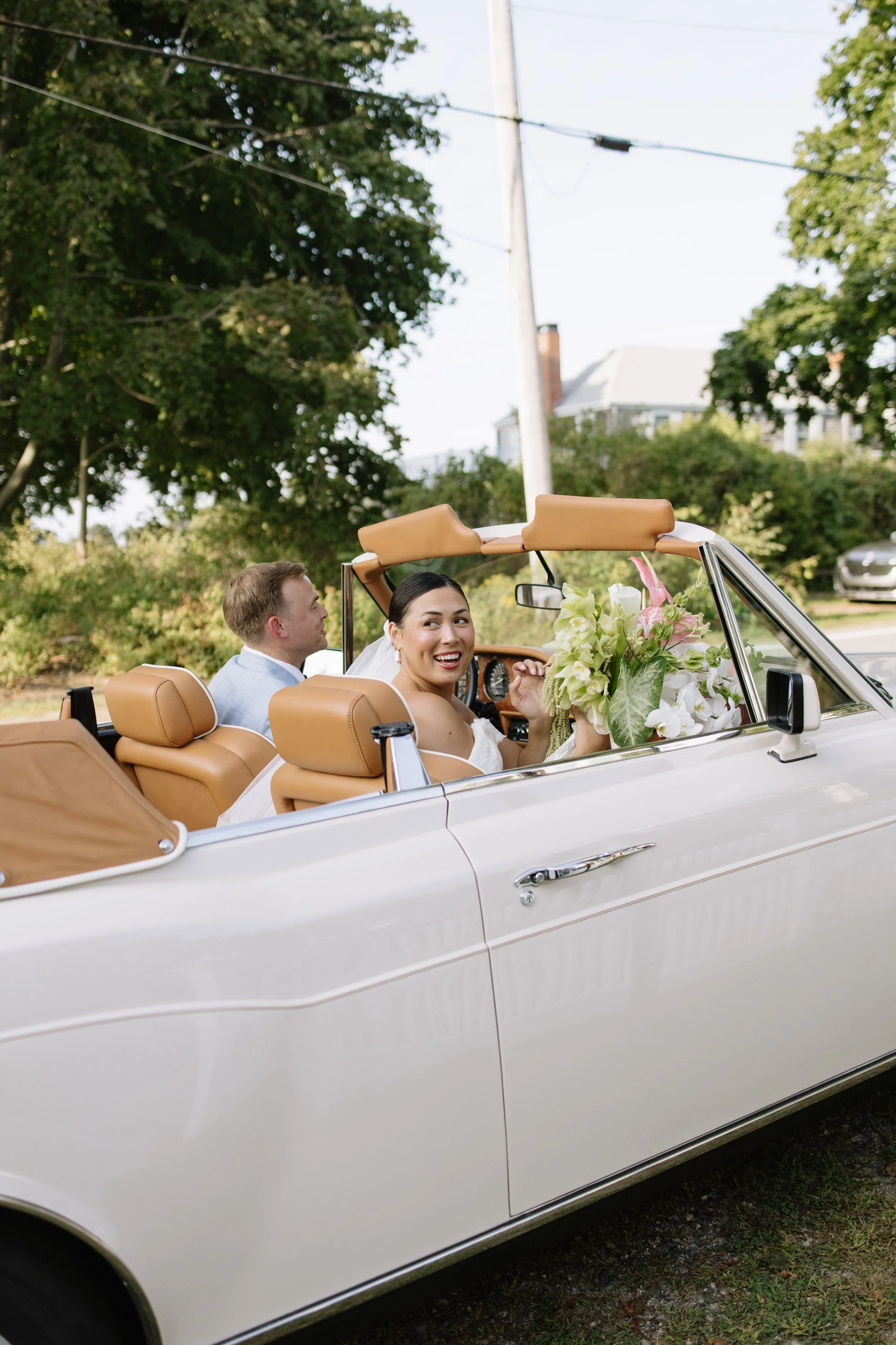 Bride and groom in a vintage convertible car with a bouquet of flowers, smiling and looking back, outdoors with trees and houses in the background.