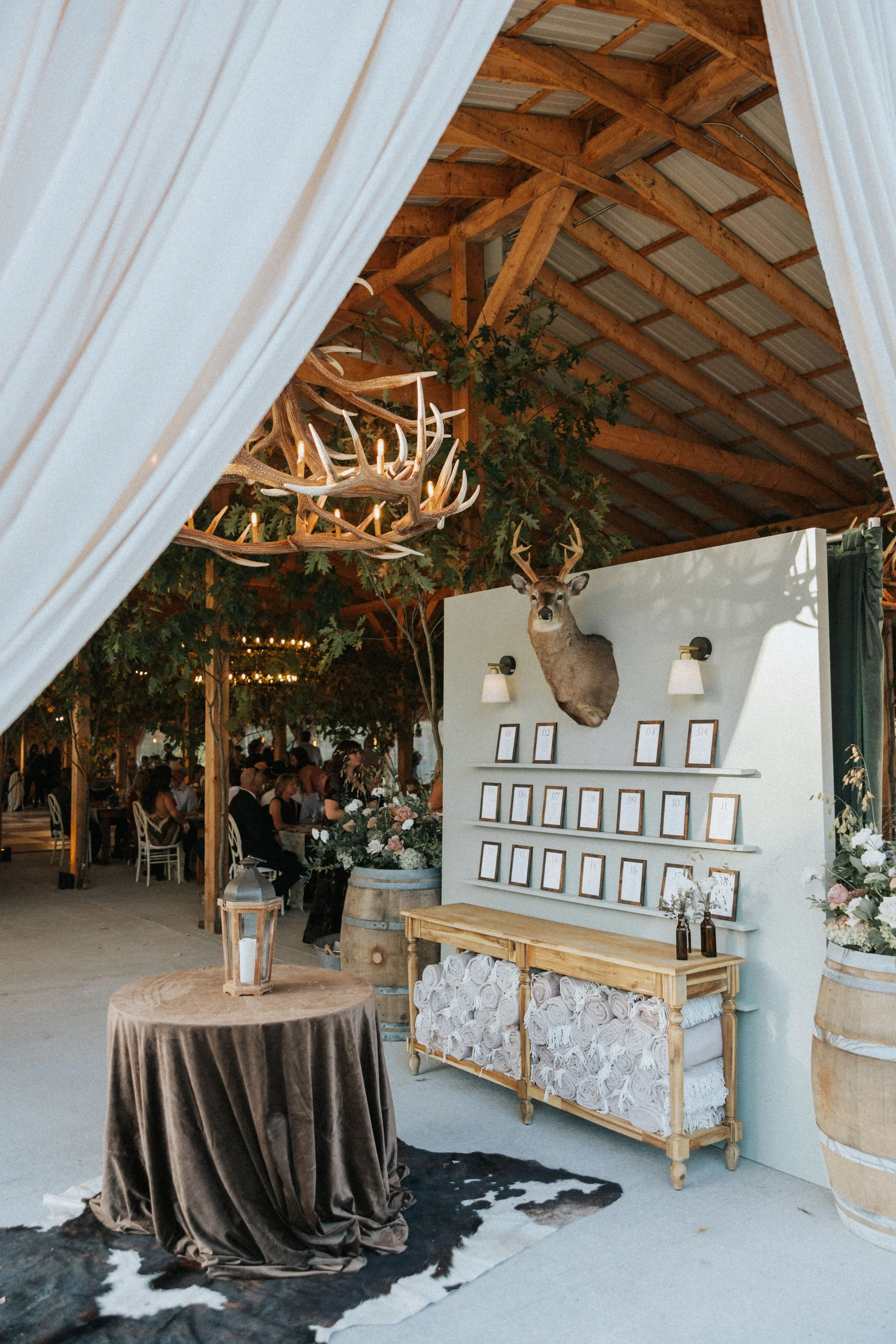 Event hall decorated with rustic and elegant decor. A white display wall features a mounted deer head and small framed items, with shelves holding more frames. To the left, a table covered with a brown velvet cloth holds a lantern, and there are re rolled towels on a wooden table underneath with decorative bottles and flowers on top. The background shows guests seated at tables with white chairs and a chandelier made of antlers hanging from the ceiling.
