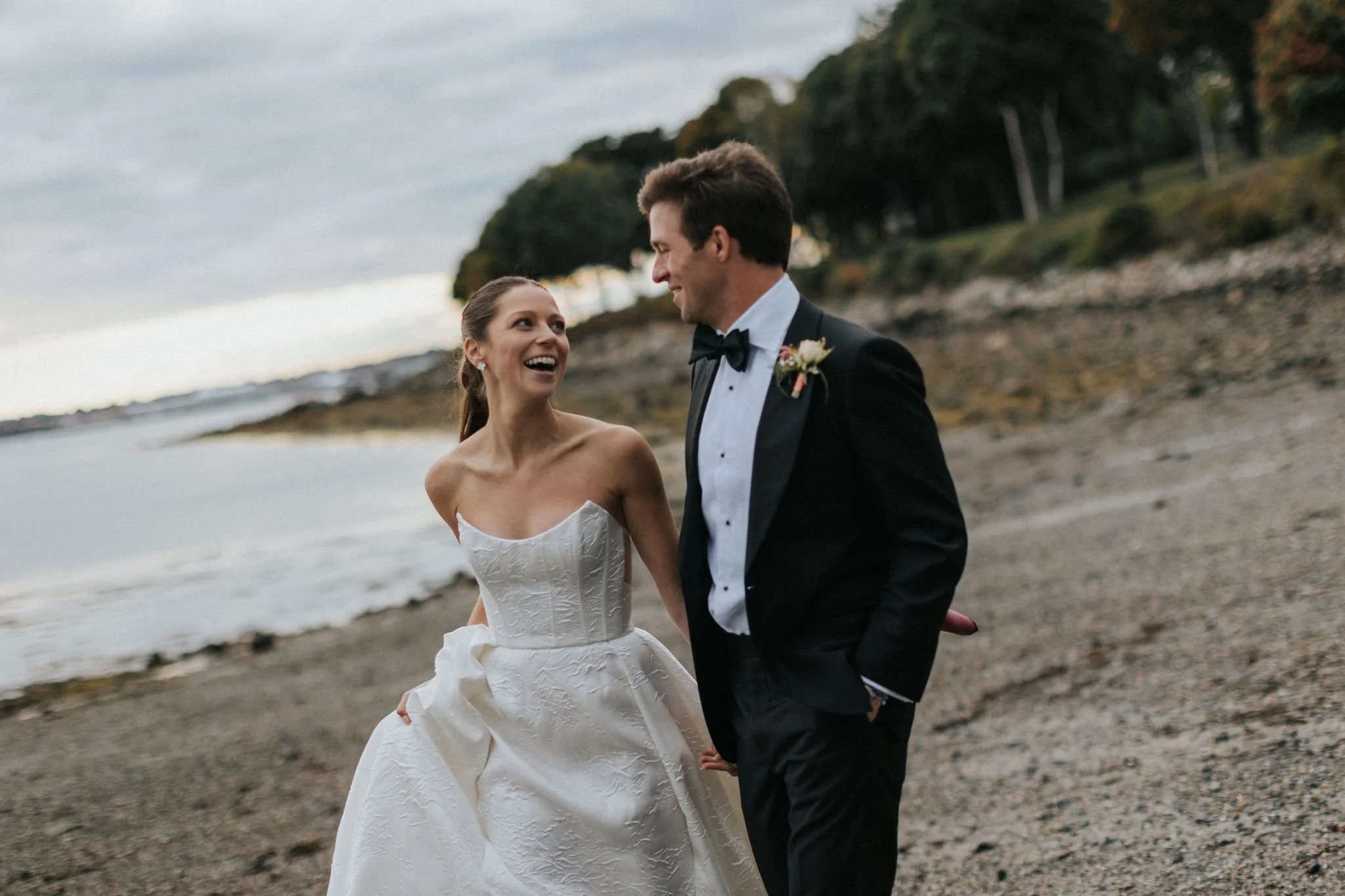 A bride and groom stand on a rocky beach, dressed in wedding attire, with the ocean and overcast sky in the background.