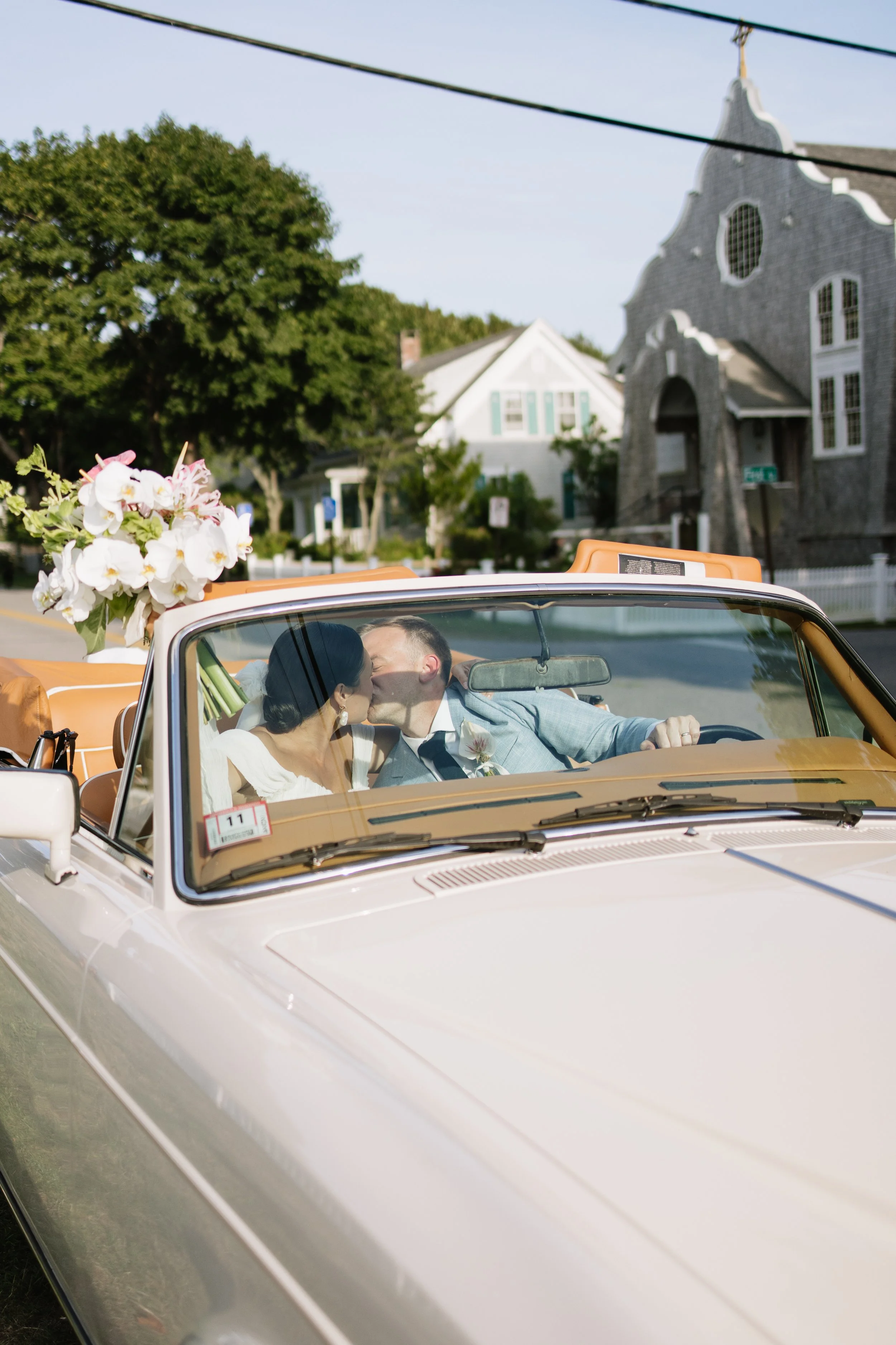 A newlywed couple sharing a kiss in a vintage convertible car decorated with flowers on the back. They are parked on a street with houses and trees in the background.