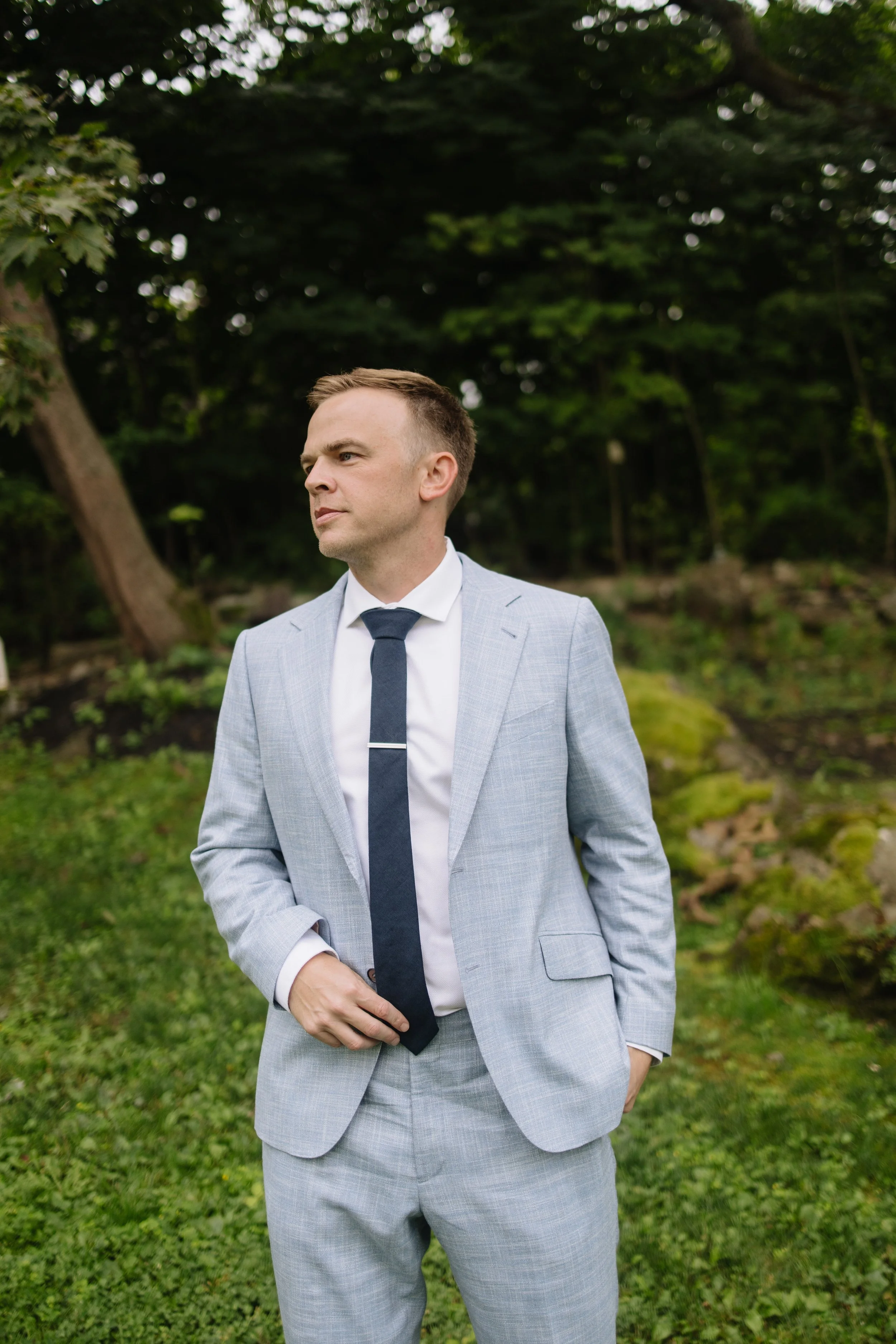 A man in a light gray suit with a dark tie stands outdoors in a lush green forested area, looking to the side with a serious expression.