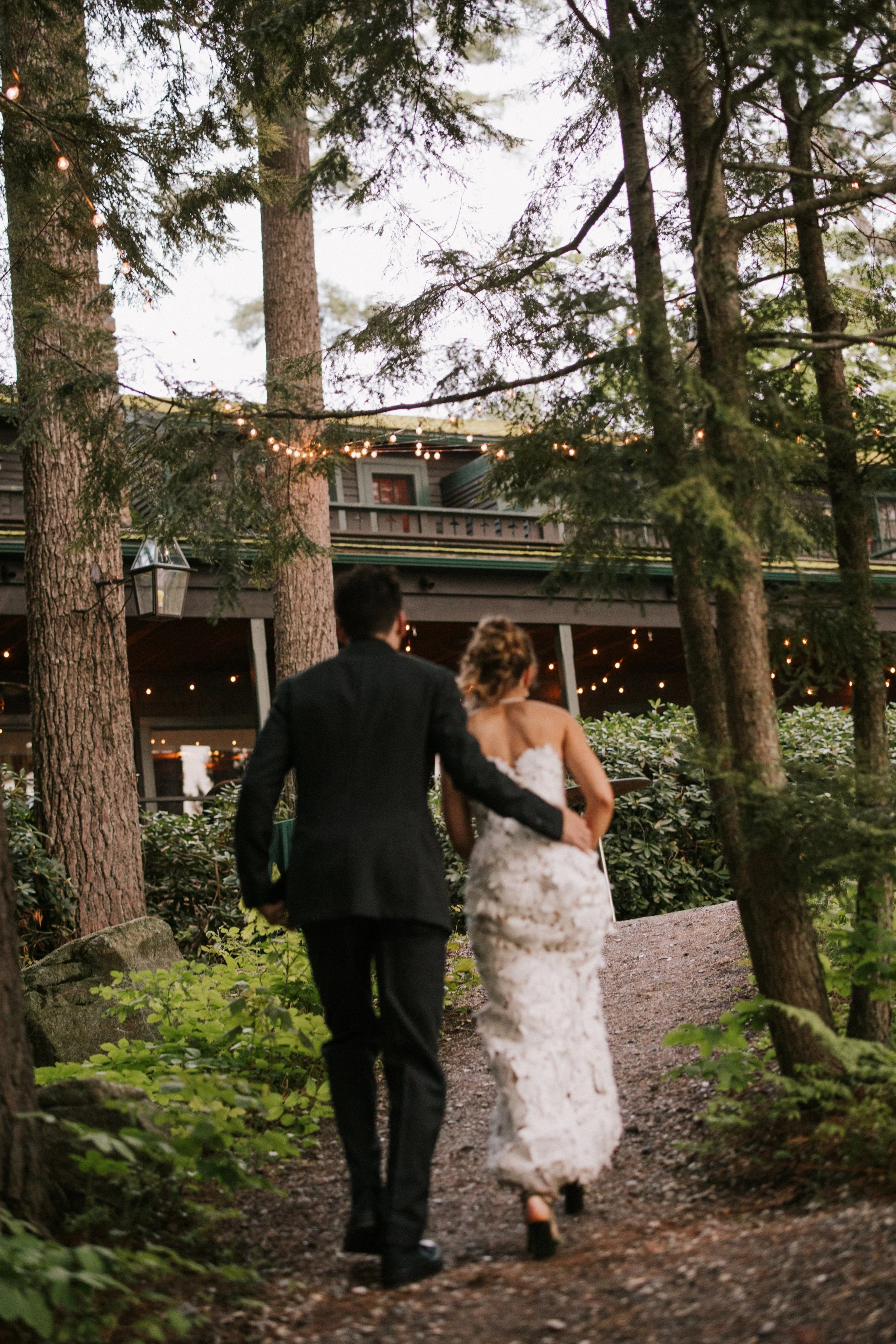 A bride and groom walking on a forest trail during an outdoor wedding at dusk, surrounded by trees and string lights.