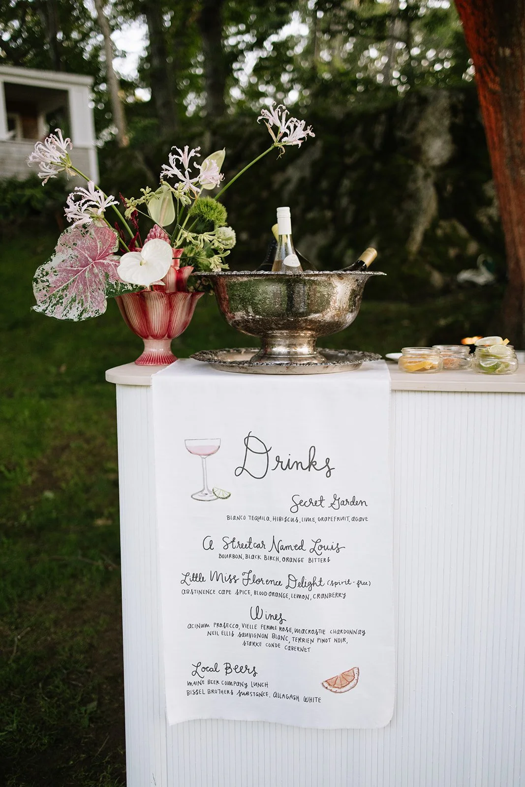 A table with a floral arrangement, a silver beverage bowl, and a drink menu sign. The menu lists various cocktails, wines, and local beers at an outdoor event during daytime in a garden setting.