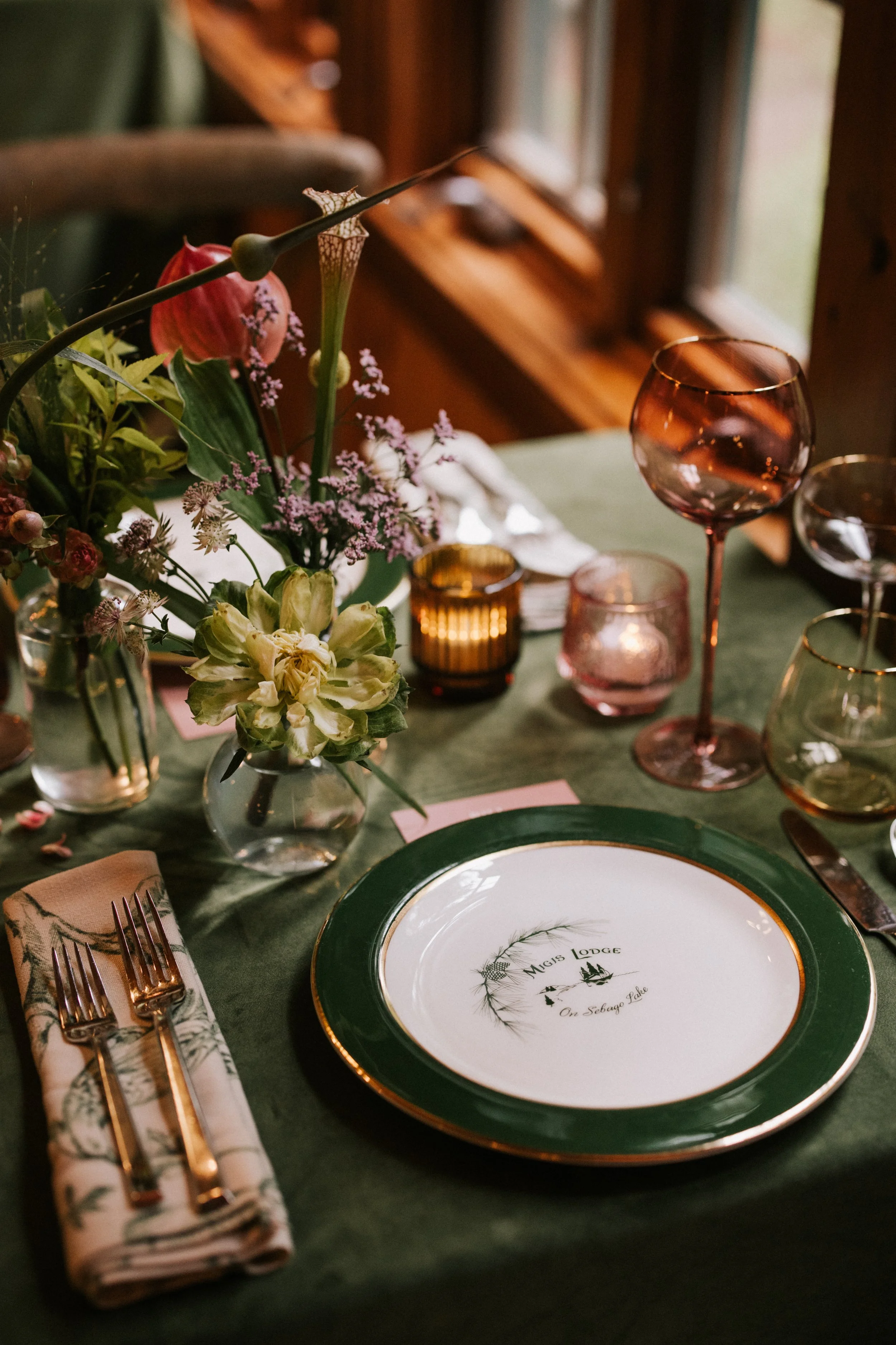 A table set for a meal with a green tablecloth, a floral centerpiece, pink and amber glassware, and a decorated plate with a gold rim and a message reading 'Mays Lodge On Sabago Lake'.