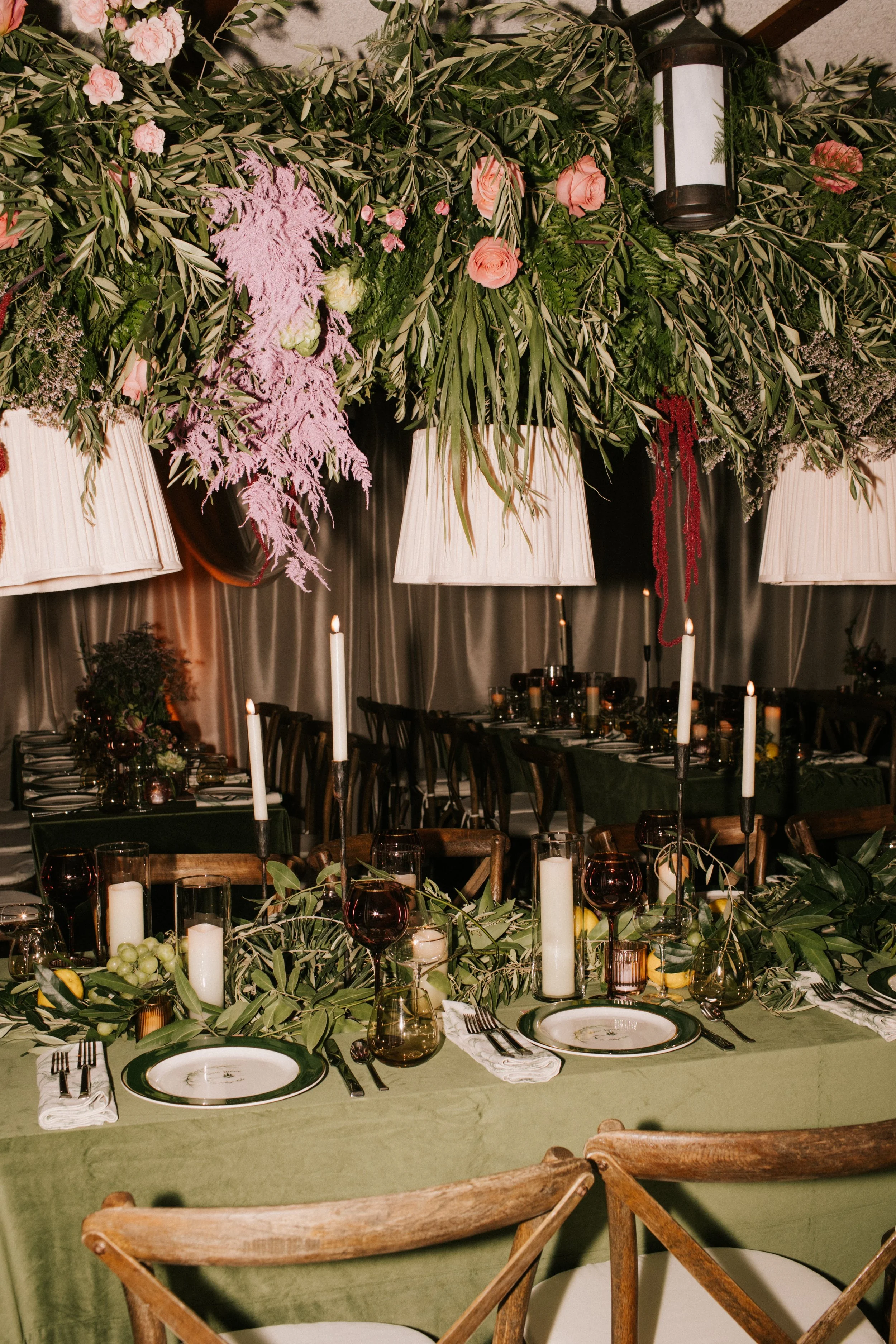 Elegant event table decorated with green foliage, candles, and dark glassware, with floral arrangements and large hanging lamps above.
