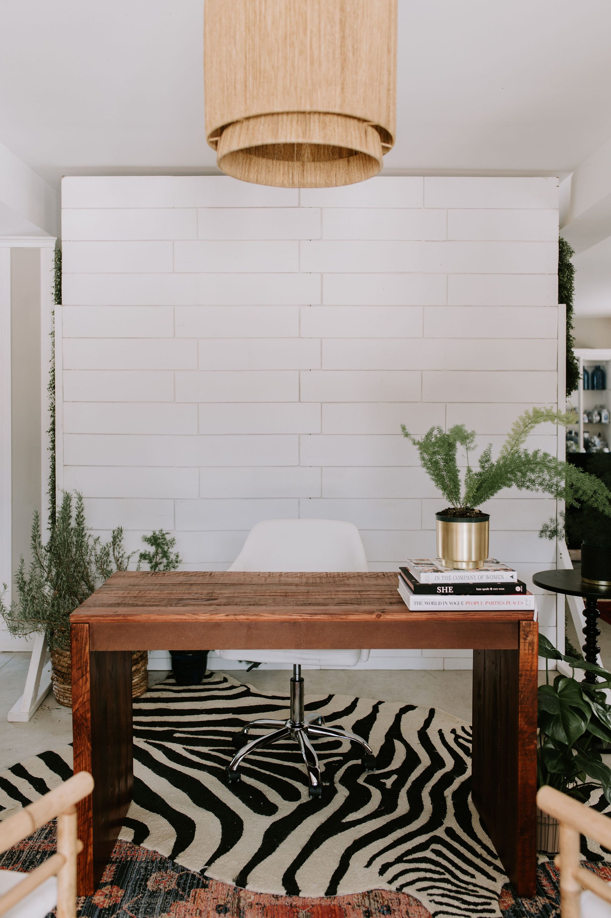 A modern home office with a wooden desk, white chair, black and white zebra rug, and potted plants, set against a white brick wall.