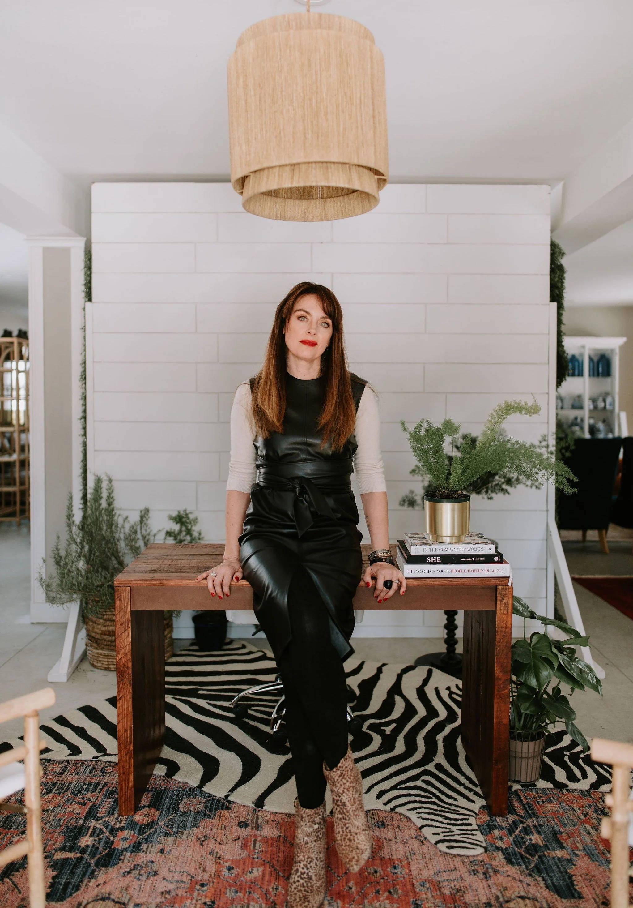 A woman with long brown hair and red lipstick sitting on a wooden table in a modern, stylish room. She is wearing a black leather dress, leopard print ankle boots, and a beige top with sleeves. The room has a white shiplap wall, plants, a striped zebra rug, and a large beige pendant light hanging from the ceiling.