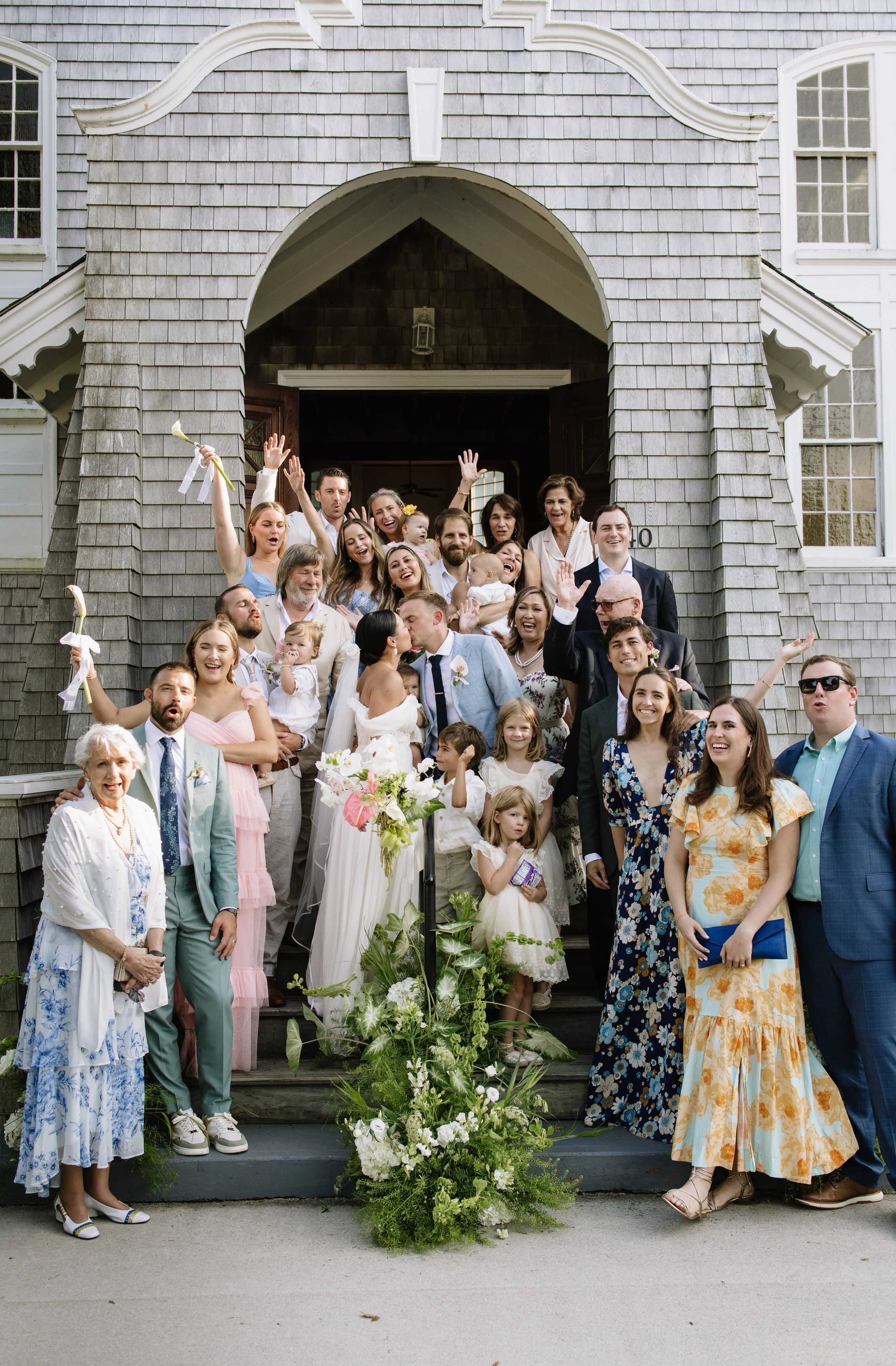 Group of people celebrating a wedding in front of a house, with a bride and groom kissing in the center, surrounded by family and friends.