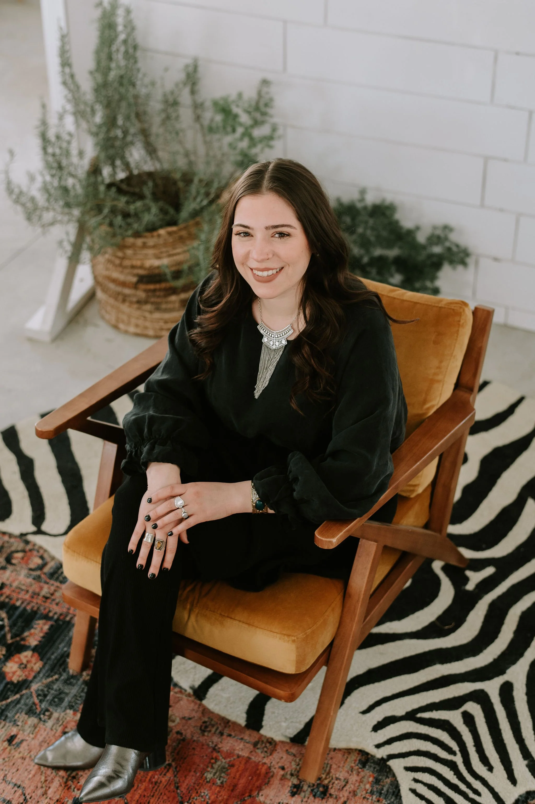 A woman with long dark hair, wearing a black outfit and jewelry, sitting in a wooden chair with a mustard-colored cushion, smiling, with potted plants and a white brick wall in the background.