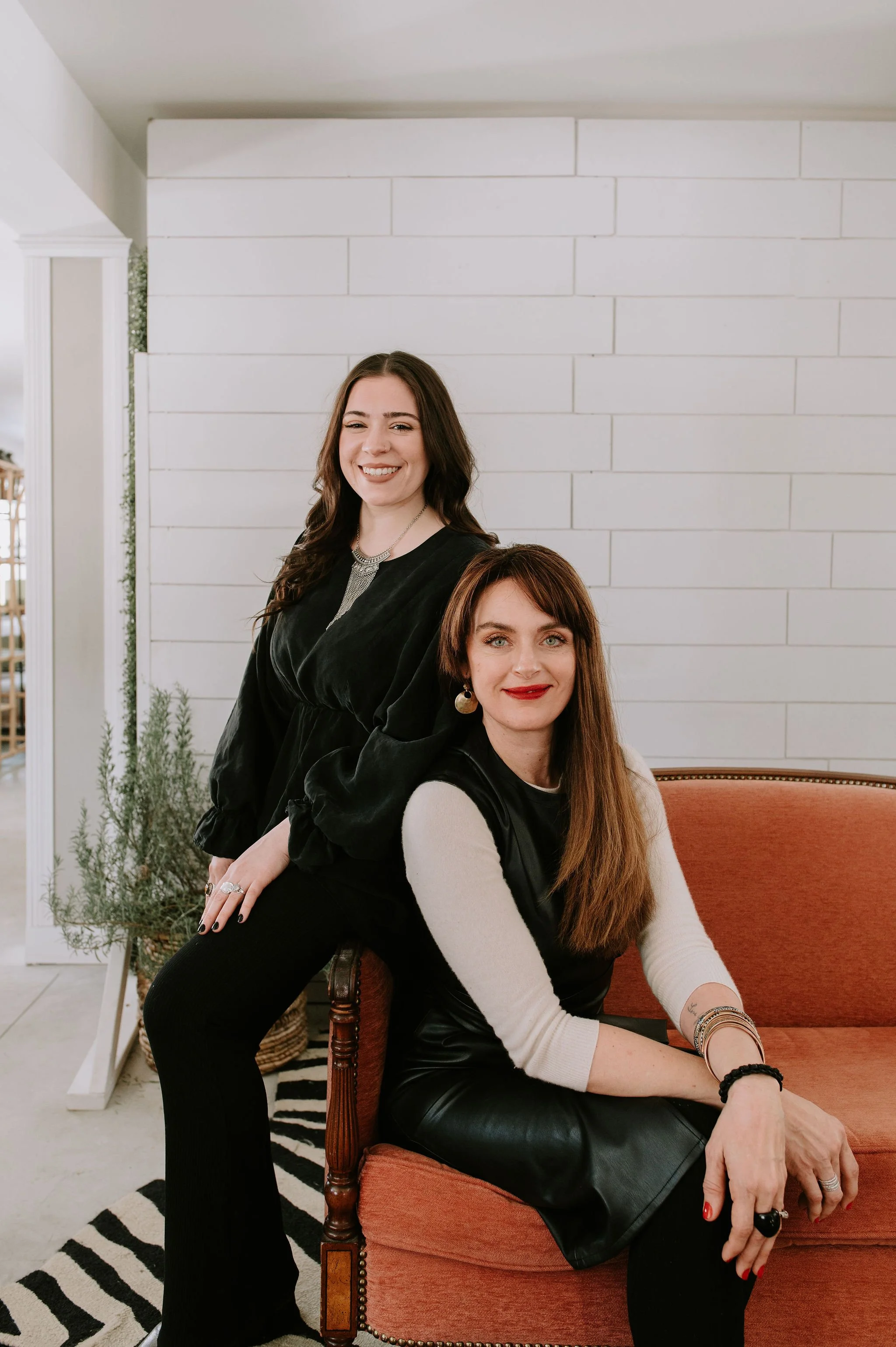 Two women posing indoors; one sitting on a red upholstered chair, and the other standing slightly behind and to the side of her. They are both smiling and dressed in stylish outfits with jewelry, with a white wall and some plants in the background.