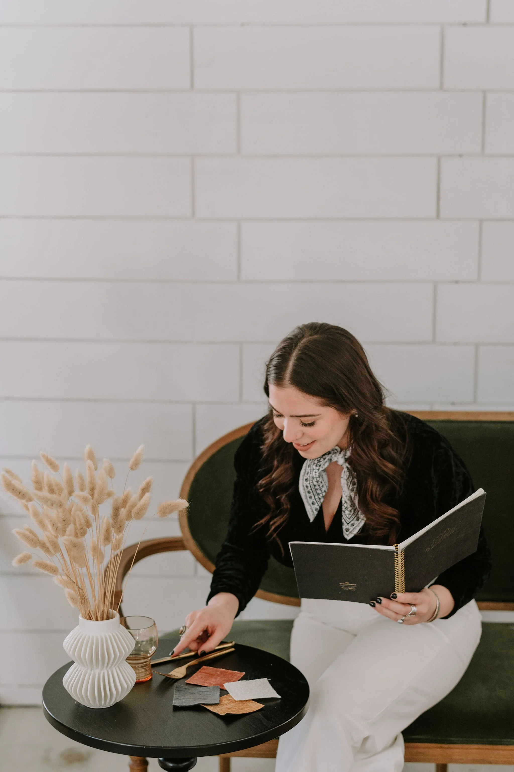 A woman sitting on a green vintage-style chair, holding a black notebook, reaching towards a small black table with color swatches, a glass, and a white vase with dried beige plants, against a white brick wall.