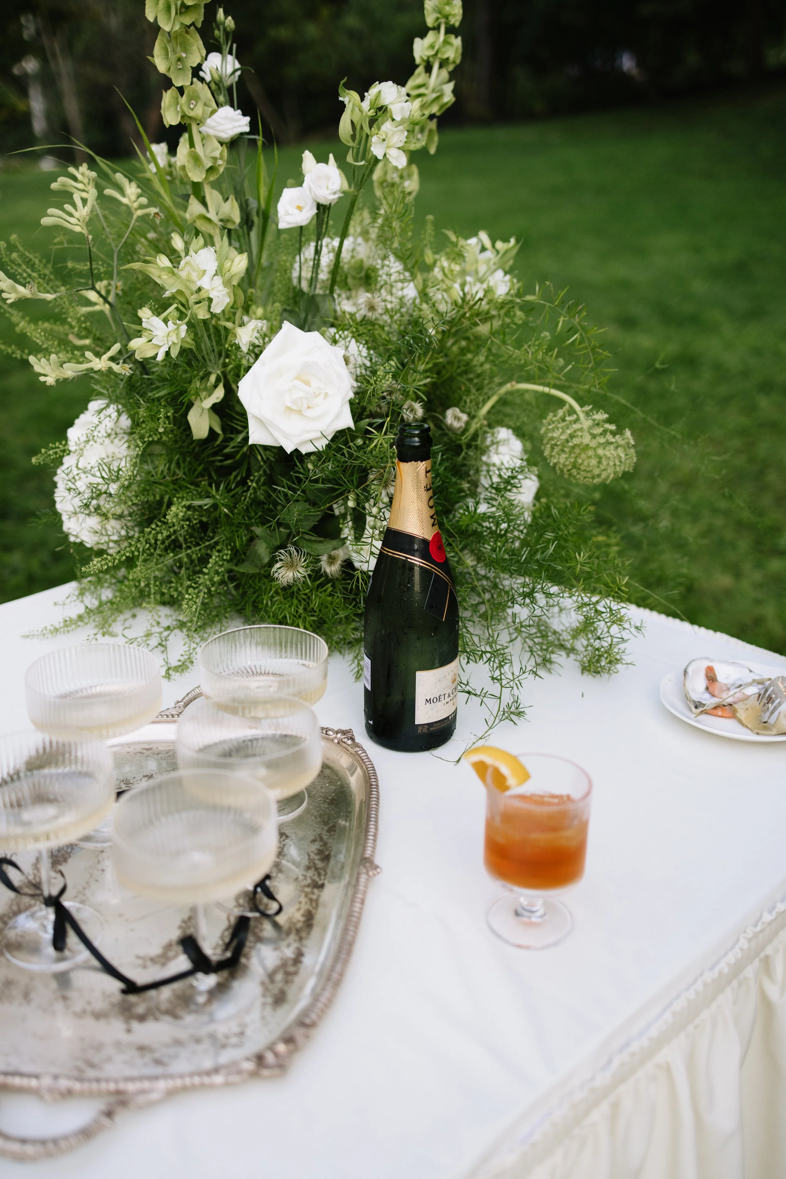 Elegant outdoor table setting with a large floral arrangement of white and green flowers, a bottle of Moët & Chandon champagne, a glass of cocktail with an orange slice, and a silver tray holding five glasses filled with clear drinks, on a white tabl