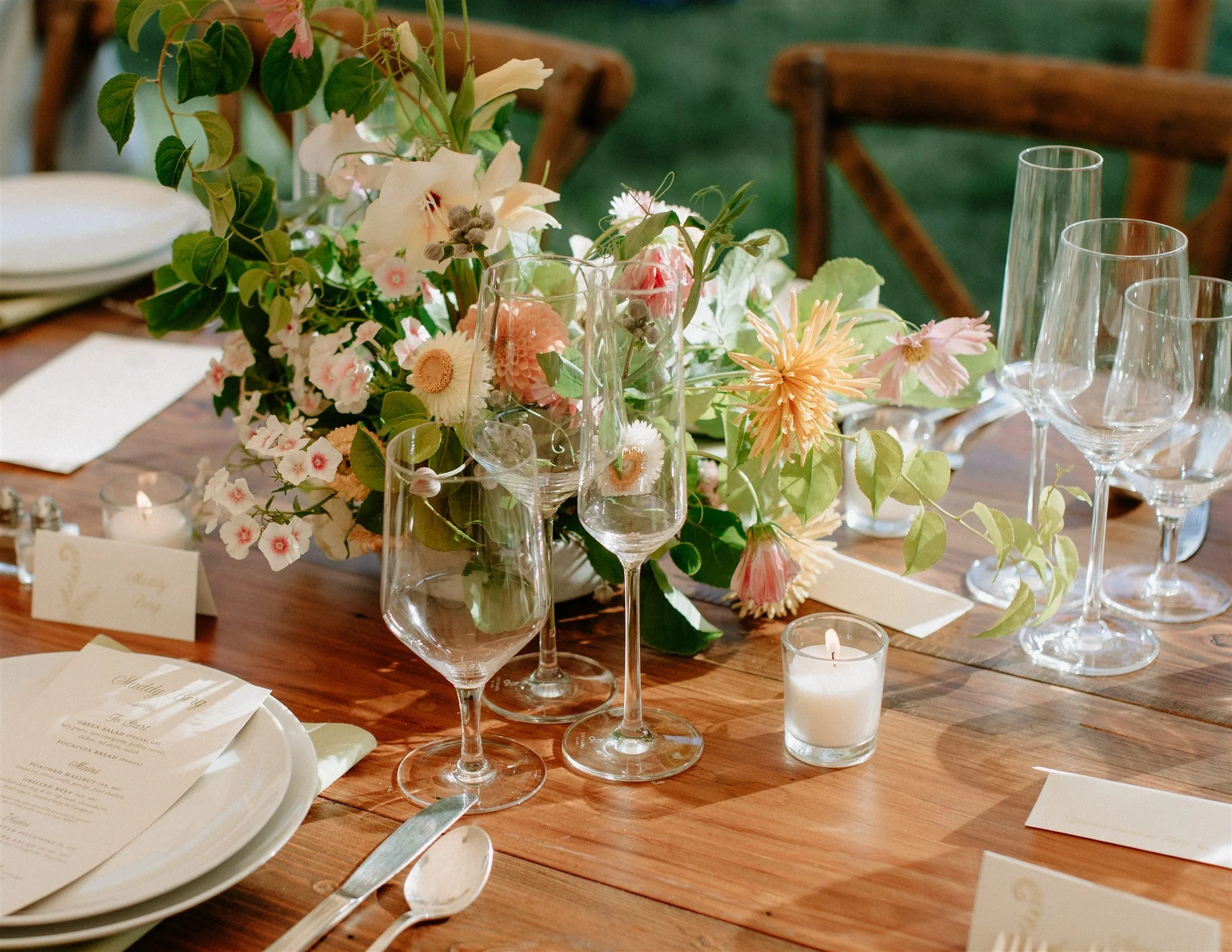 A wooden table decorated with a floral arrangement, empty wine glasses, candles, and place settings for a formal event.