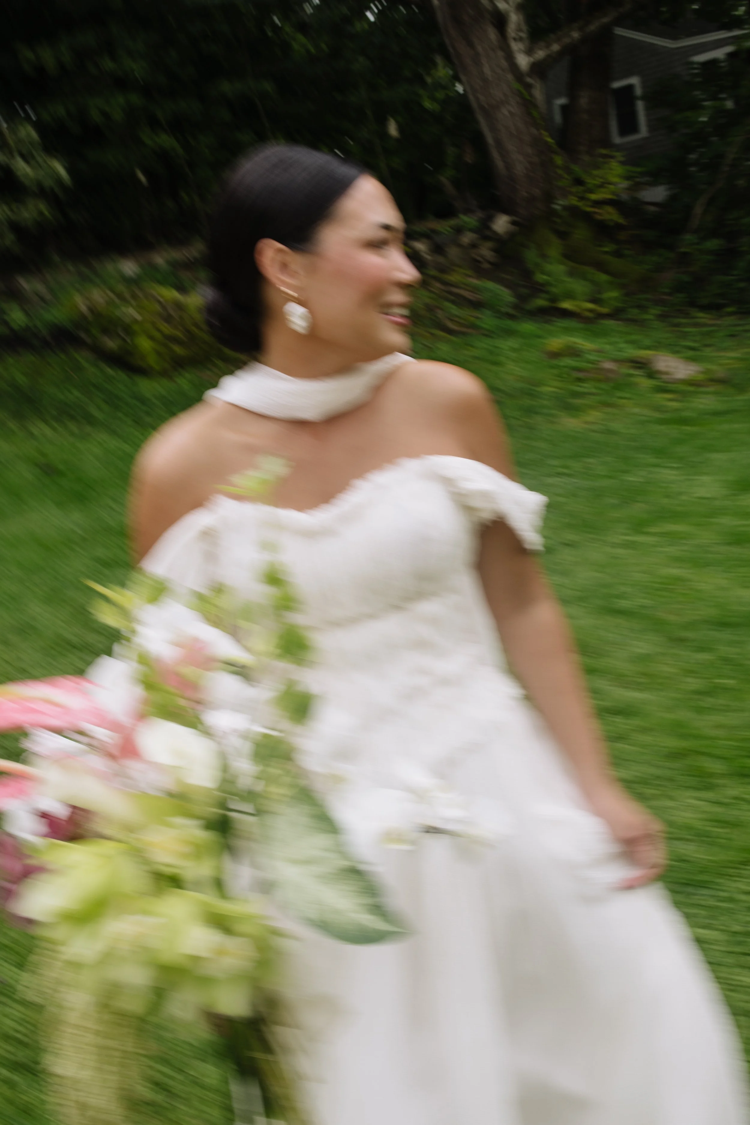 A woman in a white wedding dress holding a bouquet of flowers, outdoors with a blurred background of trees and grass.