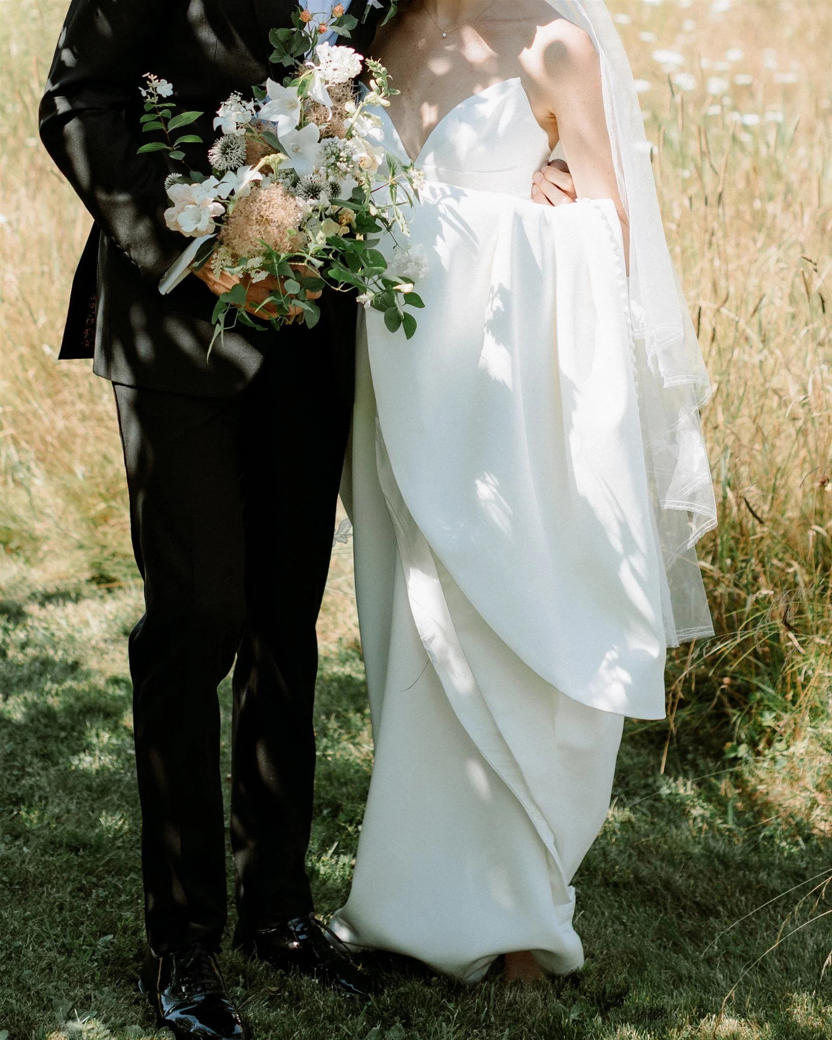 A bride and groom standing outdoors in a grassy area, with the bride in a white wedding dress holding a bouquet of flowers and the groom in a black suit.