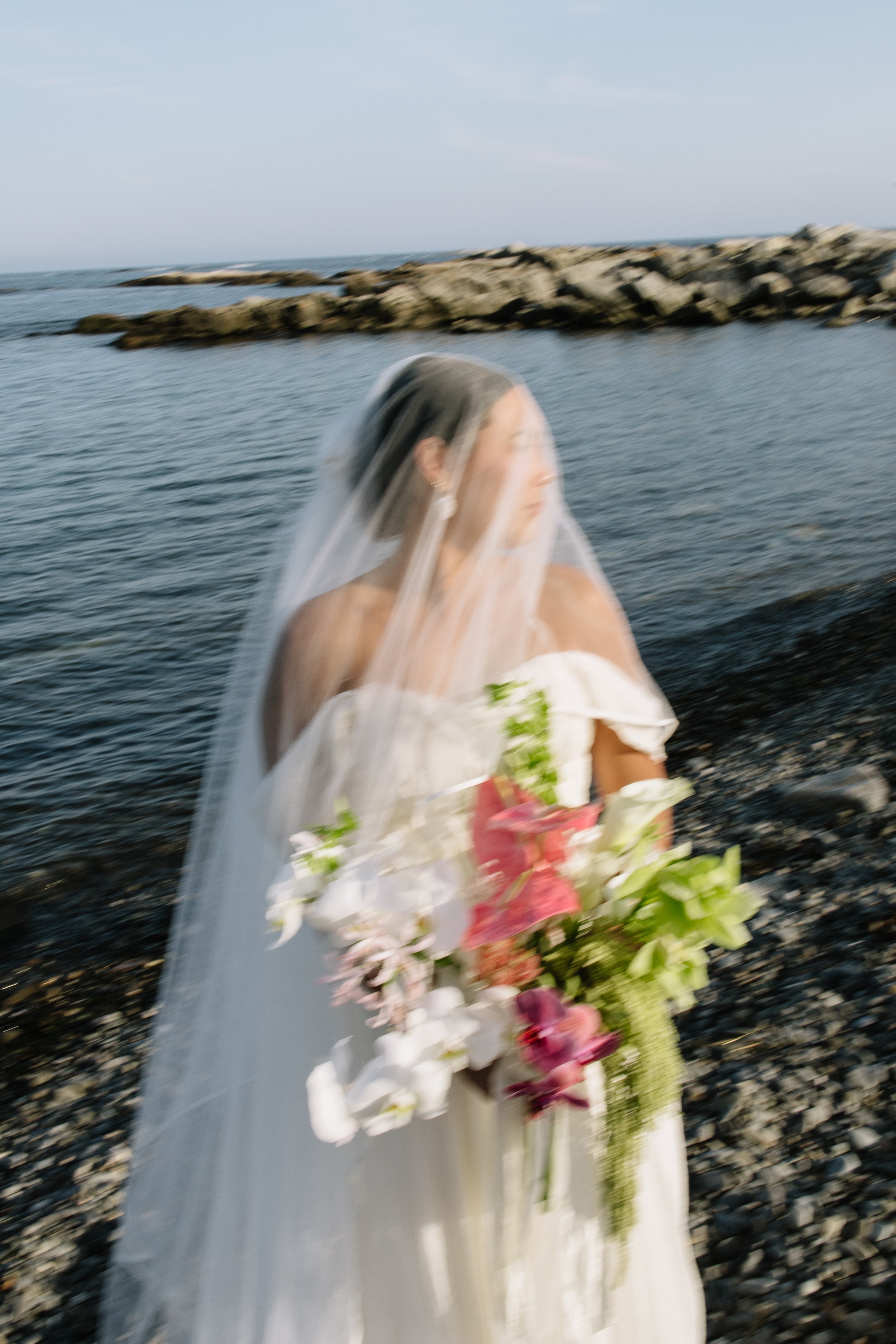 Blurry image of a bride in a white dress with a veil on a rocky shoreline, holding a bouquet of flowers, with the ocean and rocks in the background.