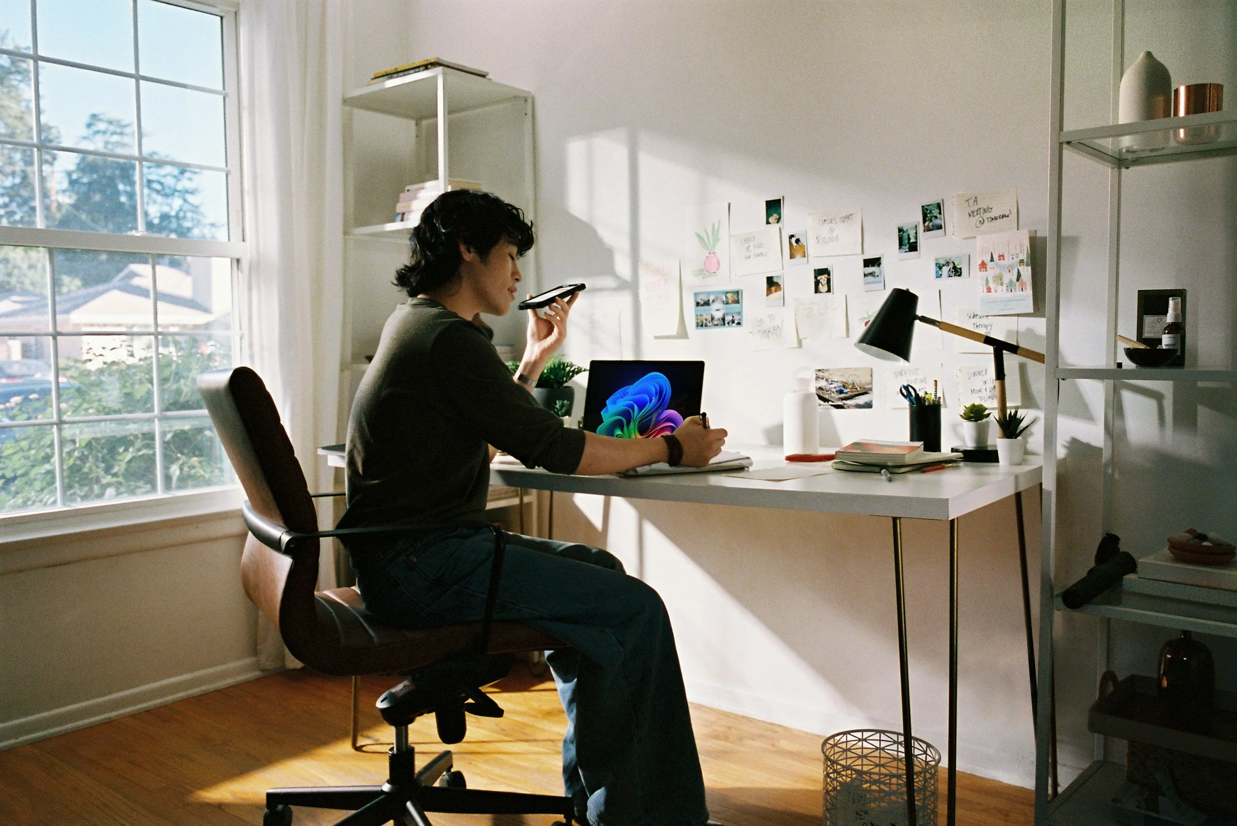 A woman sitting at a white desk talking on her cell phone while working on her computer in a sunlit home office.