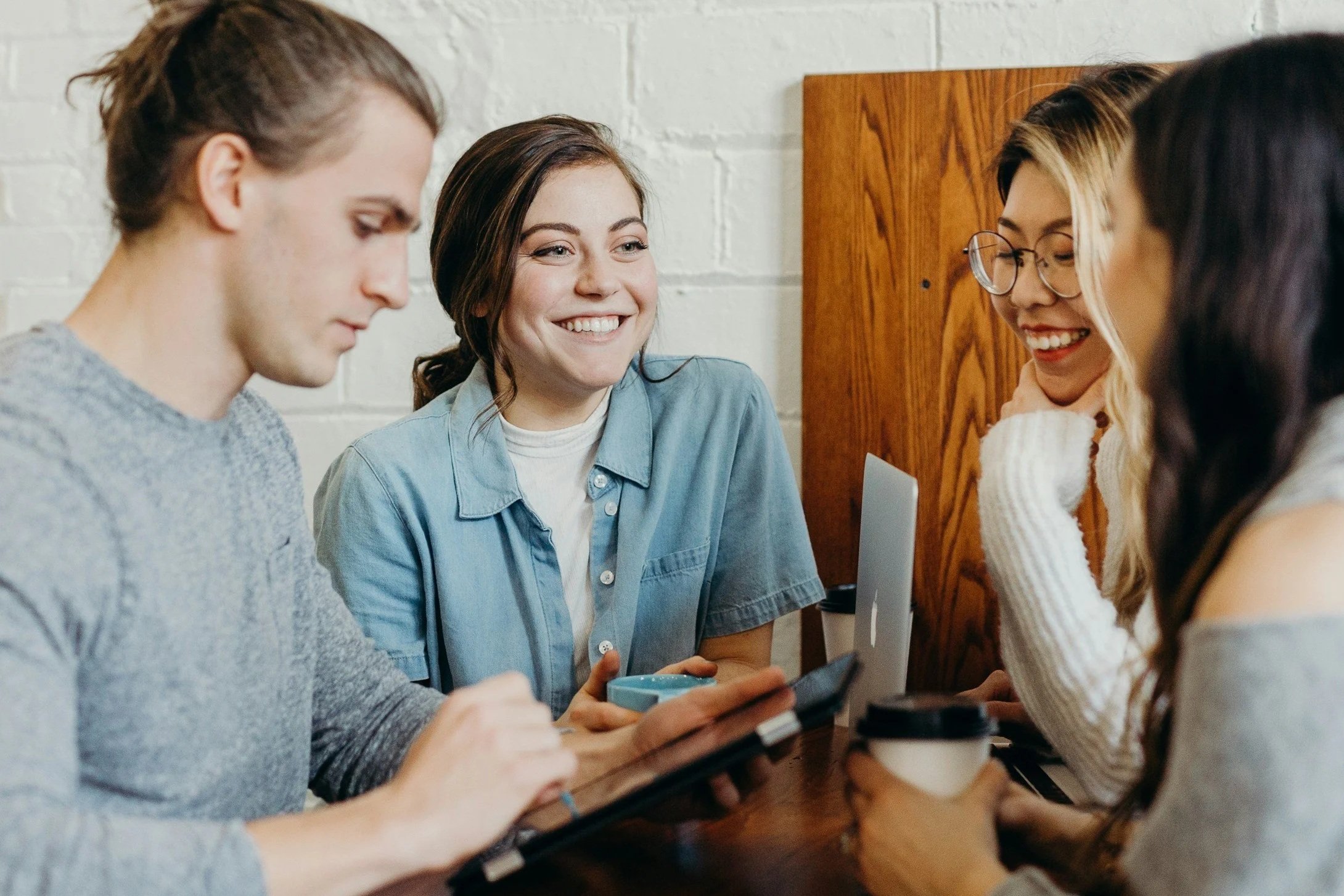 Four young adults sitting at a table, engaged in conversation and smiling, with laptops and coffee cups in a casual indoor setting.