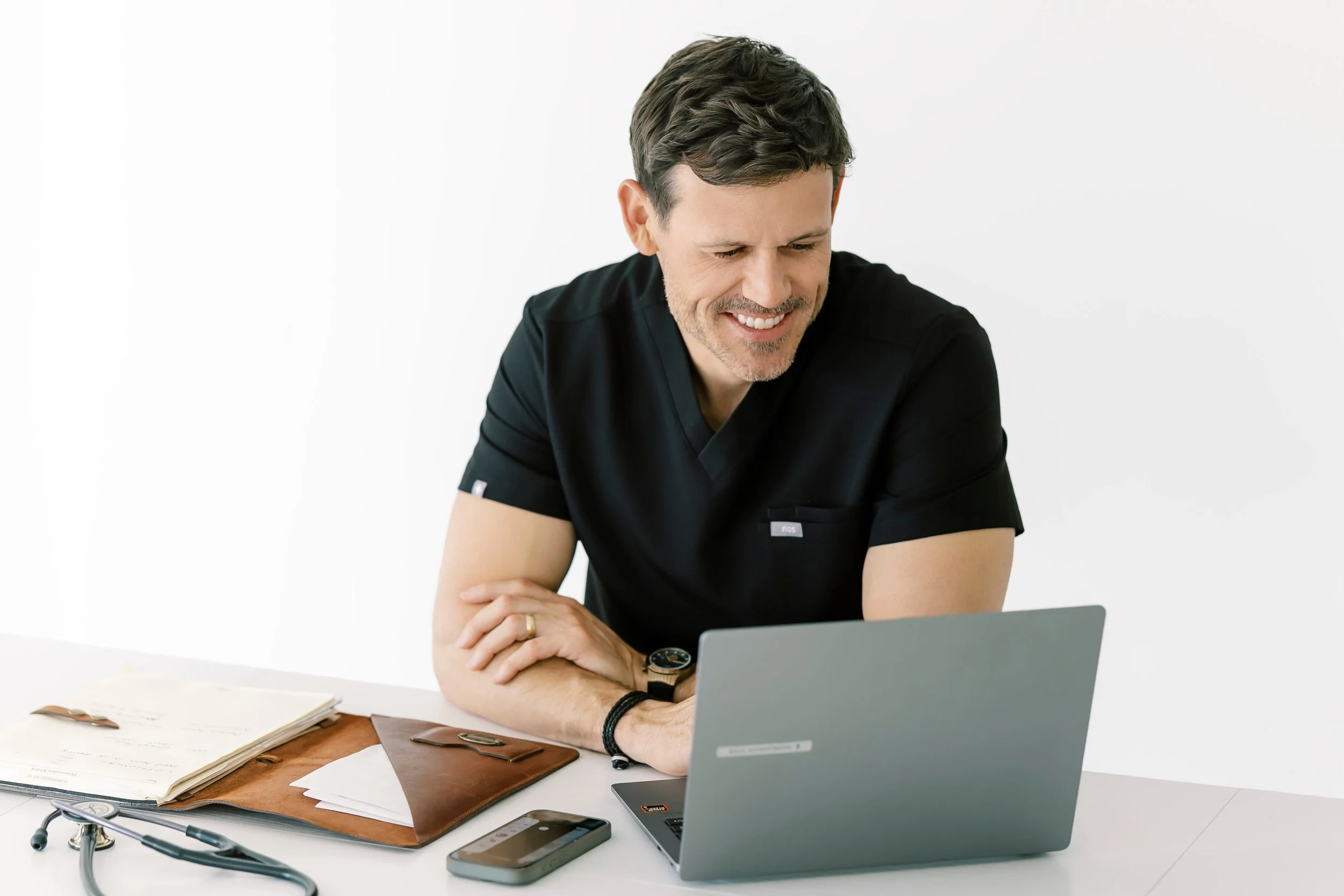 A man in a black shirt sitting at a white desk with a smile, working on a laptop. There are papers, a stethoscope, and a smartphone on the desk.