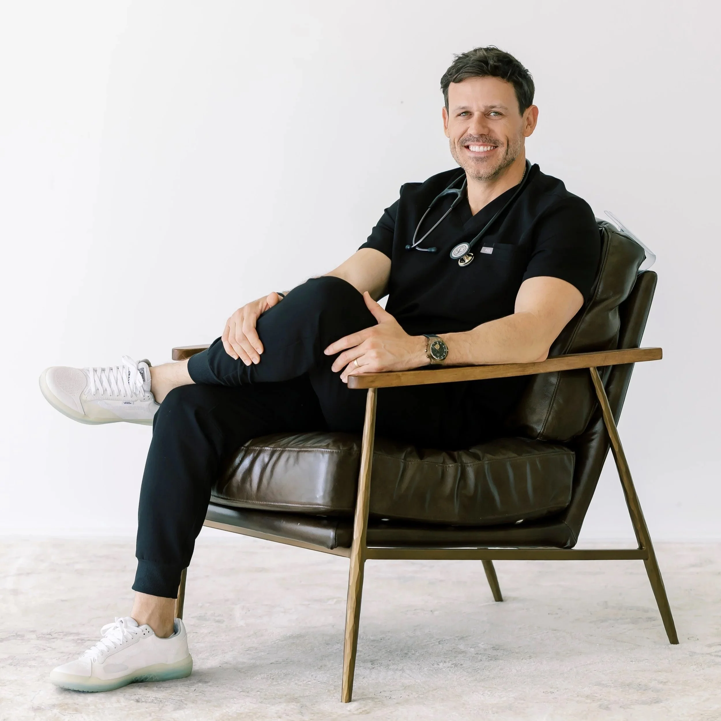 Man dressed as a healthcare worker sitting on a wooden chair with a stethoscope around his neck, smiling, wearing white sneakers and black clothing against a plain white background.