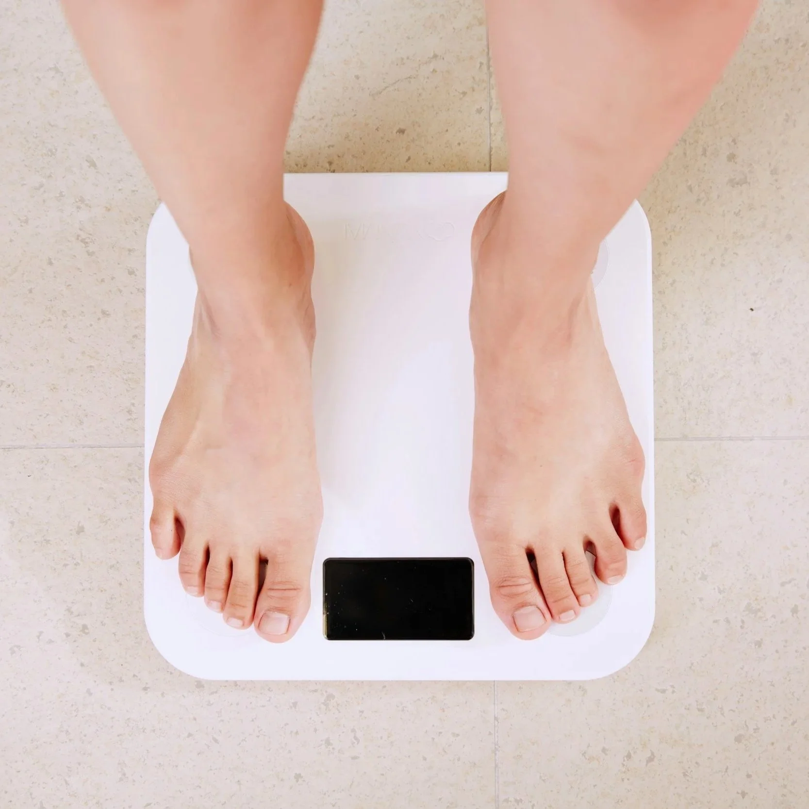 A person standing barefoot on a white digital bathroom scale, viewed from above. The scale's display is blank. The background is a light-colored tiled surface.