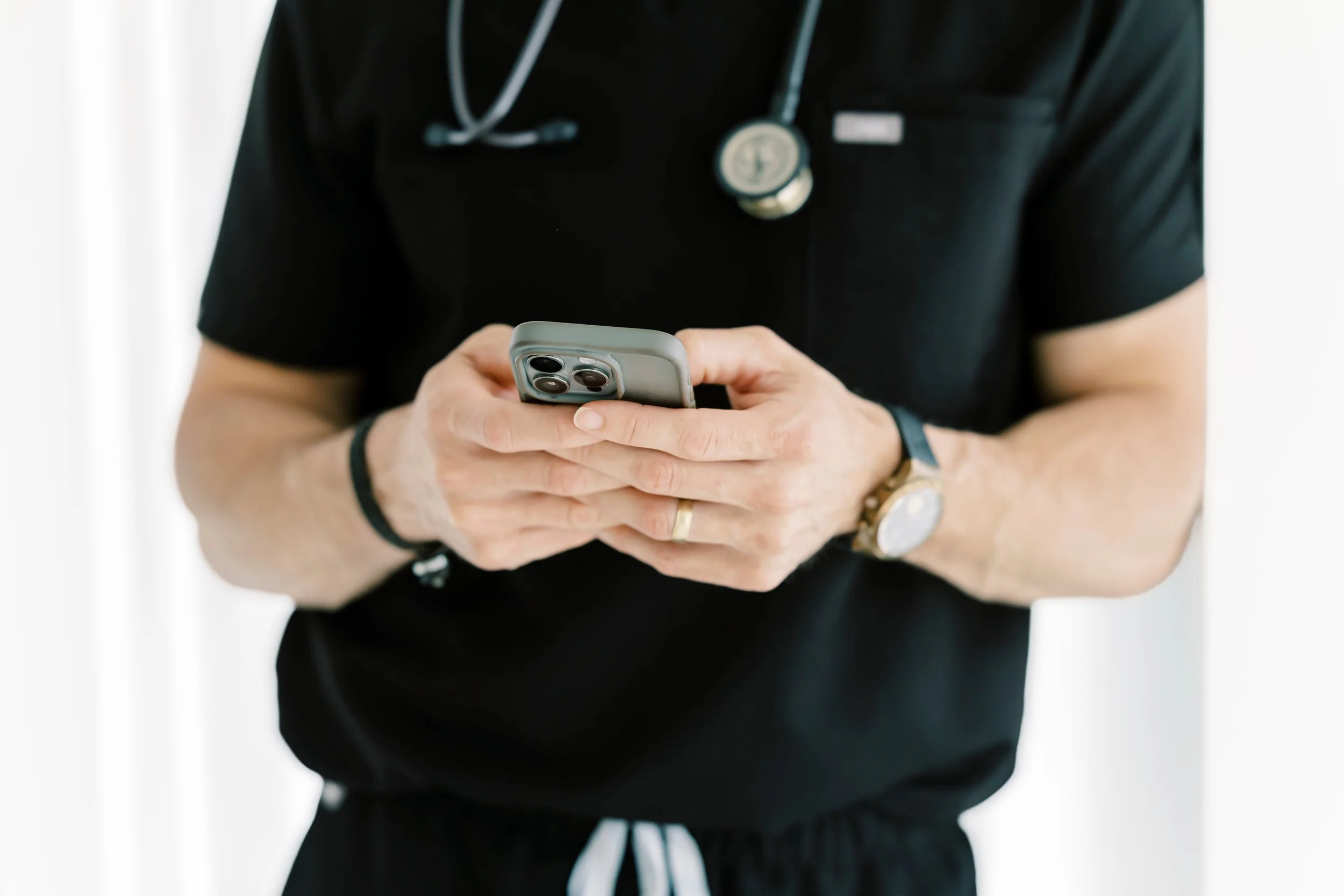 Person wearing black scrubs and stethoscope, holding a smartphone with both hands, standing against a white background.