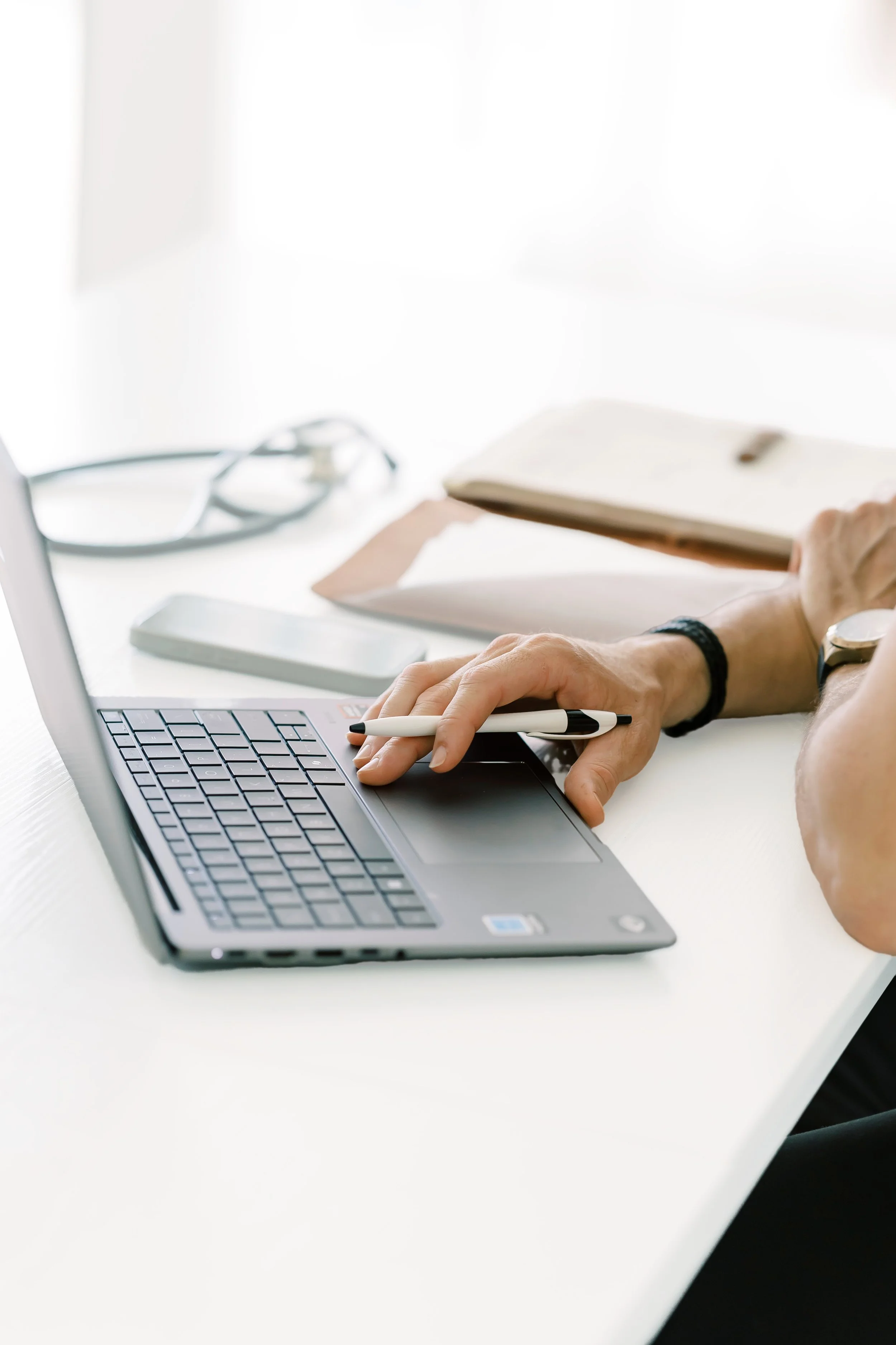 Close-up of a person using a laptop with a pen in hand, a notebook, eyeglasses, and a smartphone on a white desk.