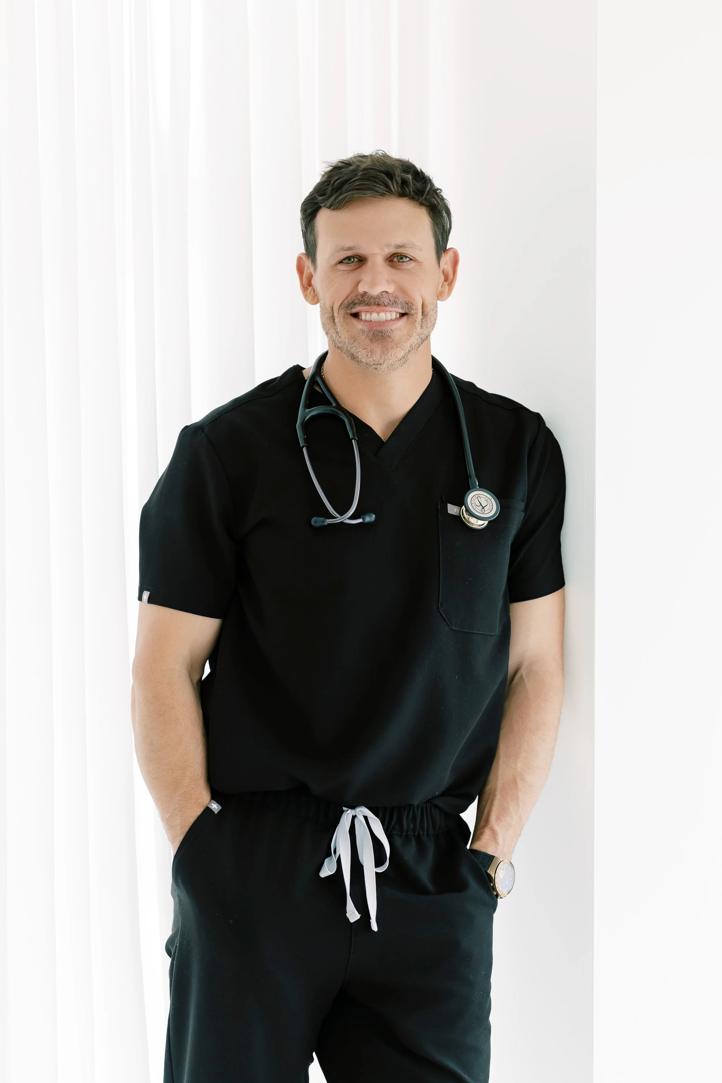 A smiling male doctor or healthcare professional with short dark hair, wearing black medical scrubs and a stethoscope around his neck, posing in front of a white background with vertical blinds.