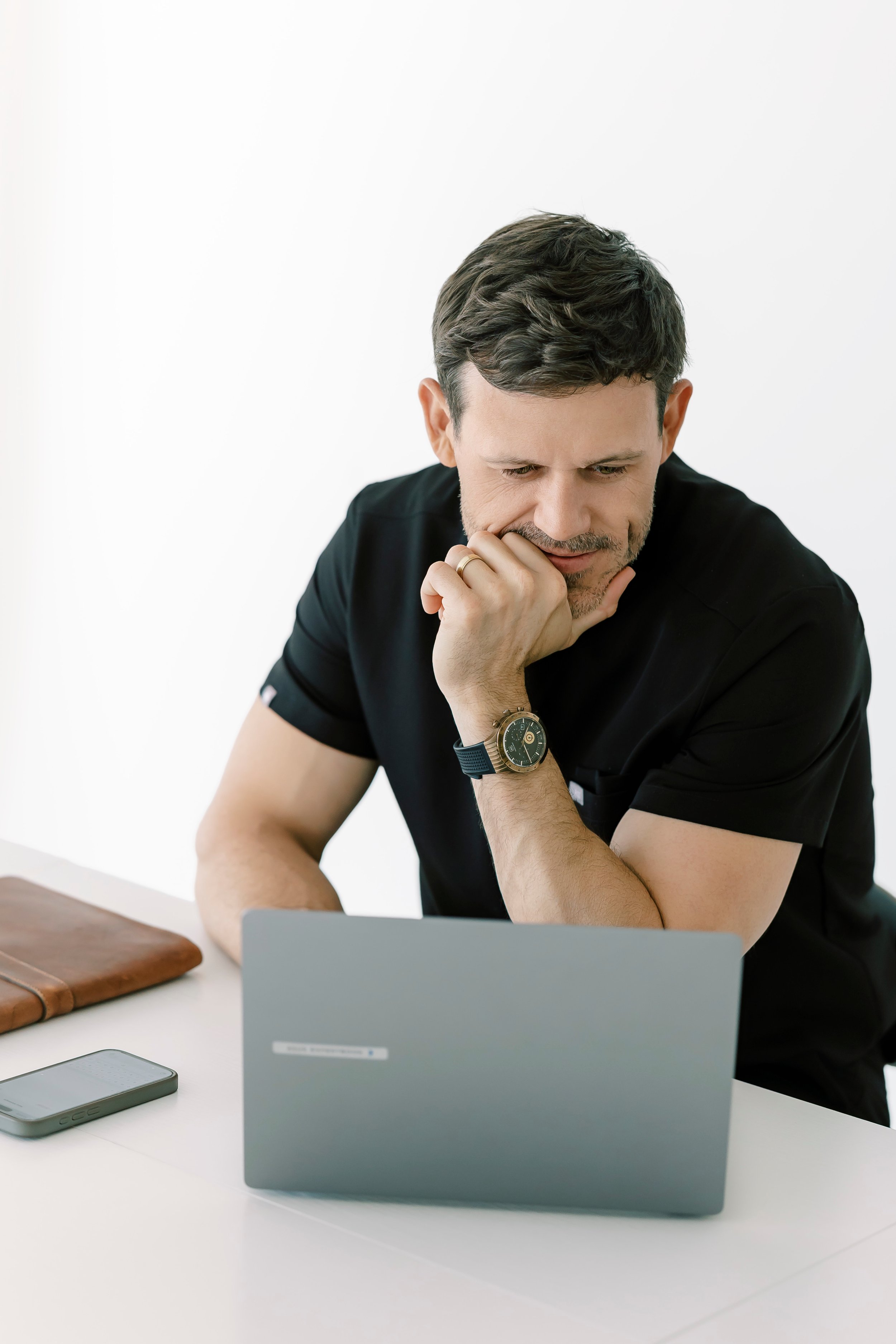 A man with dark wavy hair and a beard sitting at a white desk, looking at a laptop screen with a thoughtful expression, resting his chin on his hand. The desk has a brown leather notebook, a smartphone, and a silver tablets on it.