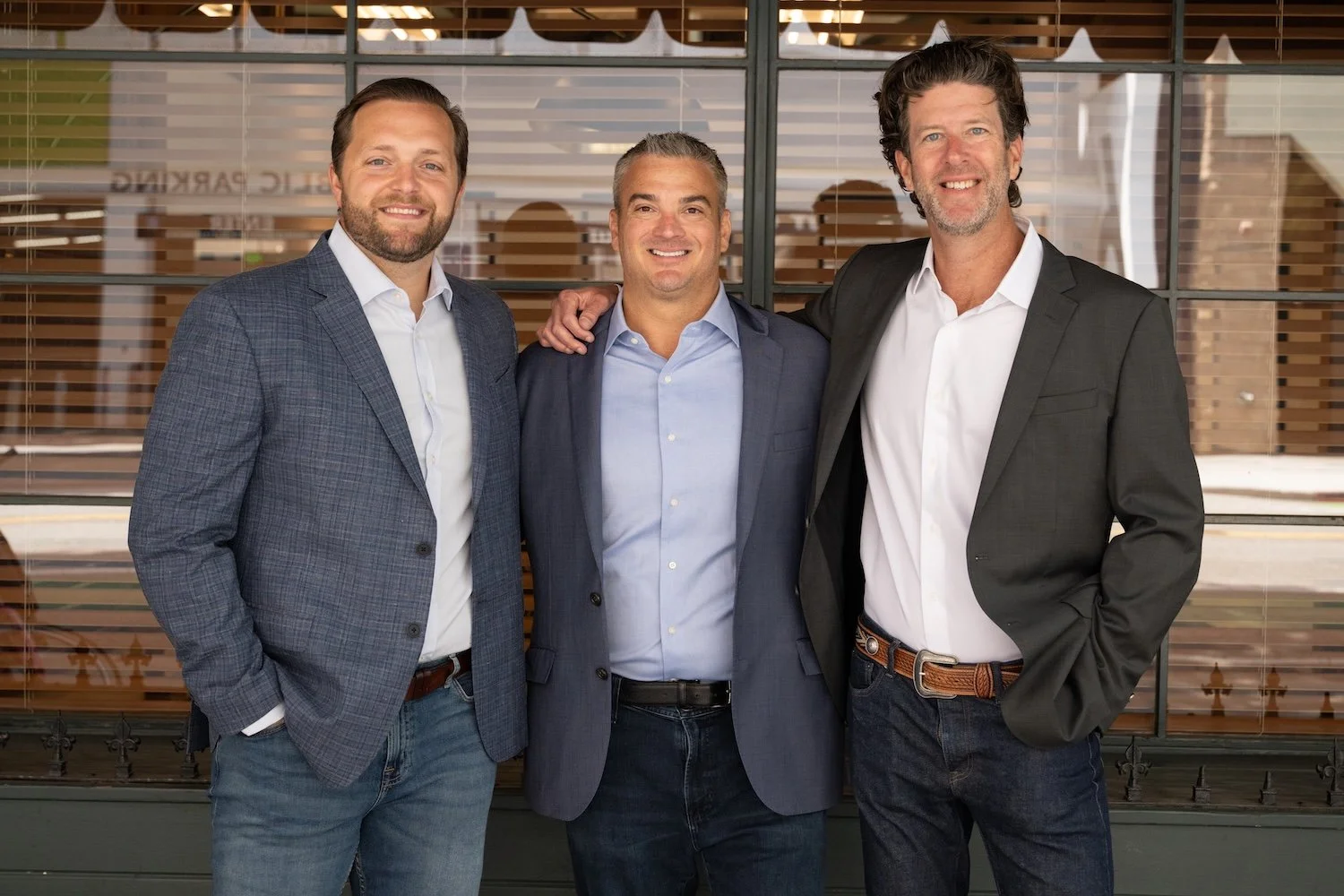Three men in business suits standing together outdoors, smiling, with glass windows and wooden blinds in the background.