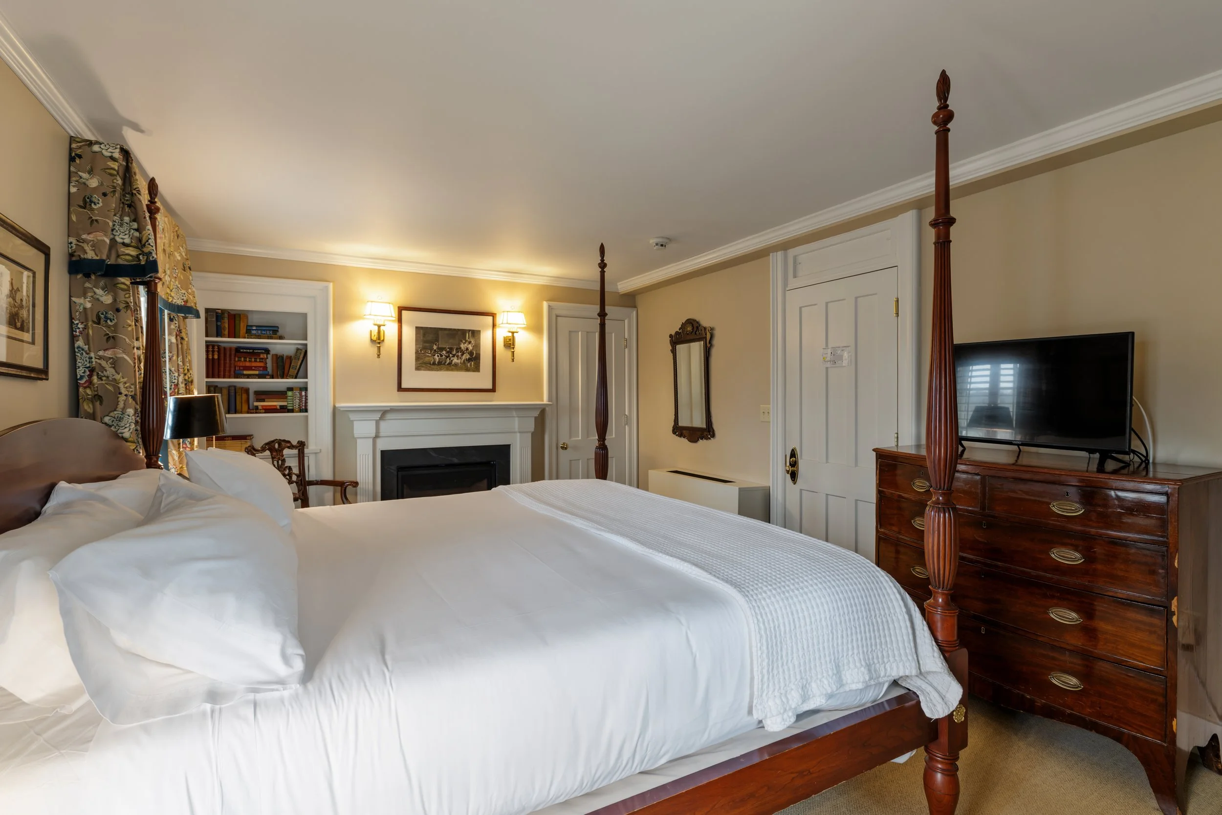 A cozy bedroom with a four-poster bed, white bedding, and pillows. There is a dresser with a flat-screen TV, a mirror on the wall, a built-in bookshelf with books, and artwork. The room has warm lighting from wall sconces, floral curtains, and beige walls.