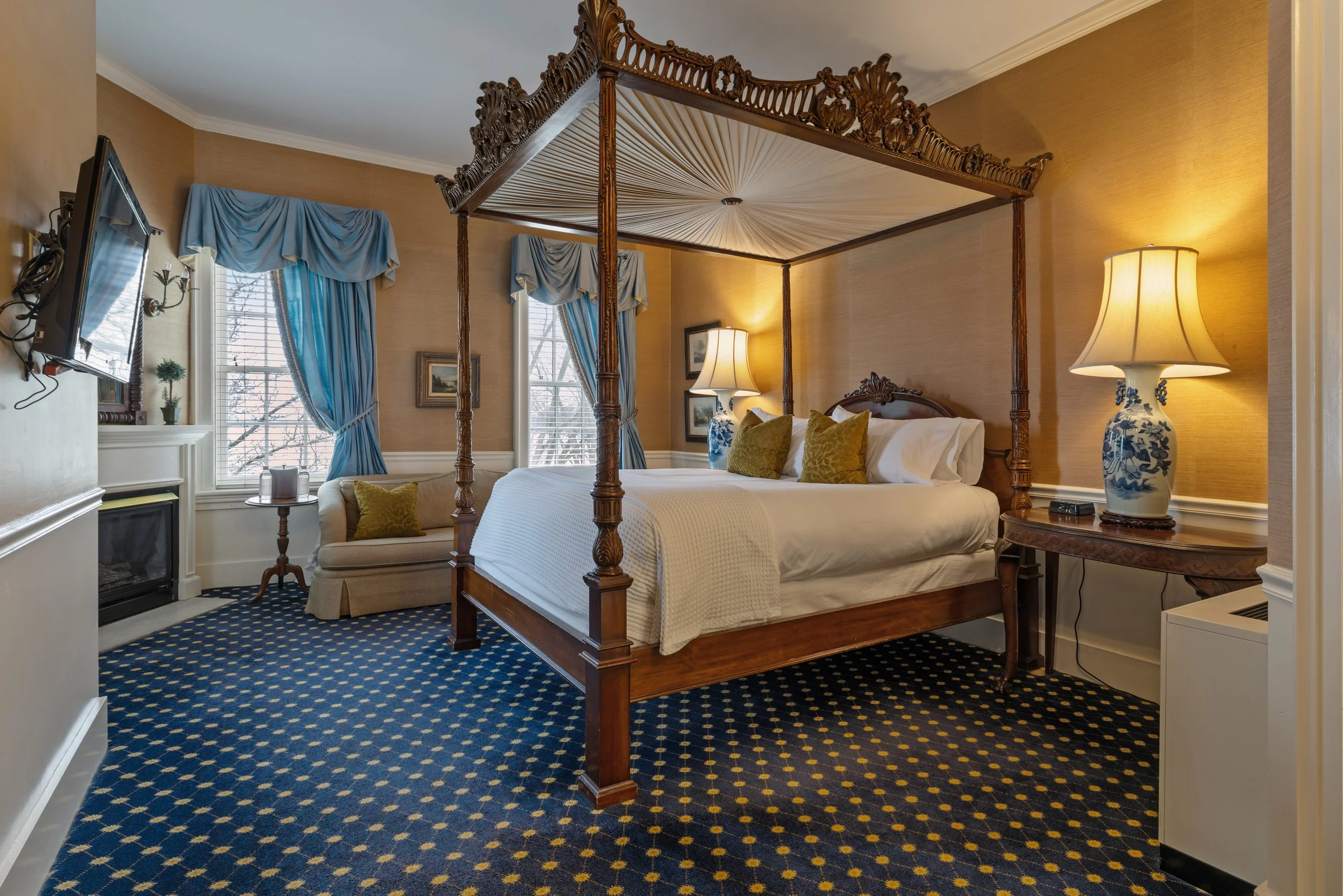 A bedroom with a four-poster bed, blue curtains, lamps, and a patterned carpet.