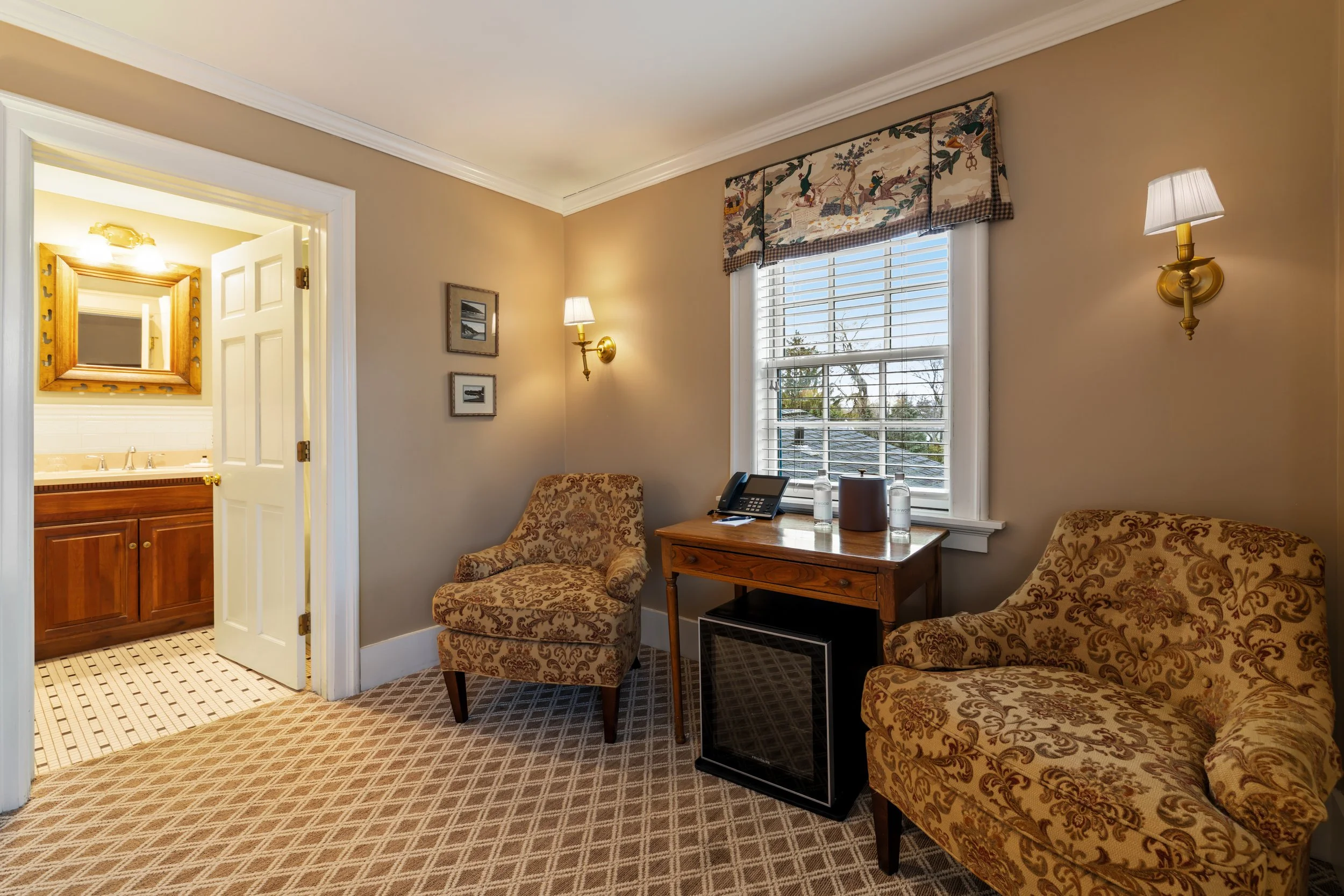 A cozy hotel room with cream-colored walls, featuring two patterned armchairs and a small wooden table with water bottles, a phone, and a speaker. There's a large window with white blinds and a decorative valance, and two wall-mounted lamps. The room extends to a bathroom with a wooden vanity and a mirror.