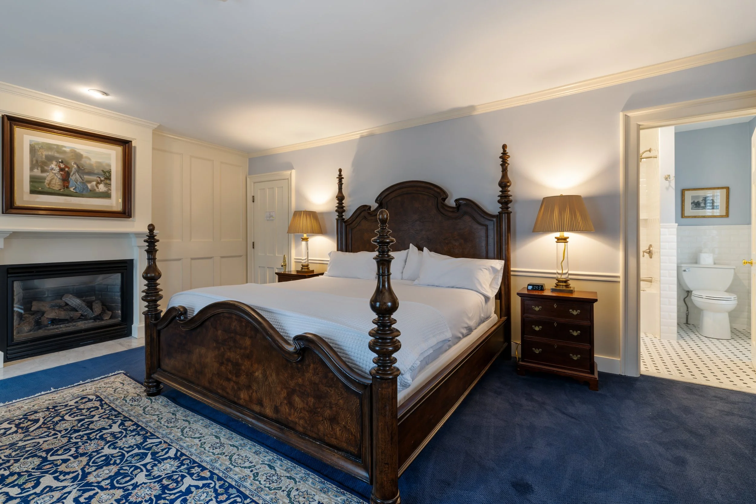 A hotel bedroom with a wooden four-poster bed, two bedside tables with lamps, a fireplace, and a bathroom visible through an open door.