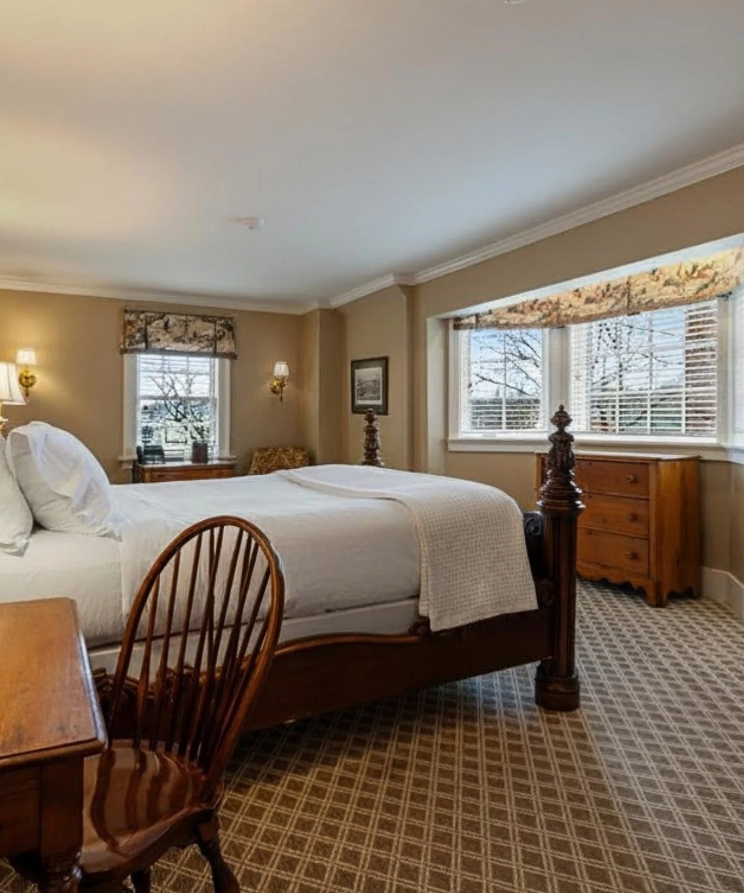 A cozy bedroom with a bed, wooden nightstands, and windows with floral valances and blinds, featuring beige walls and patterned carpet.