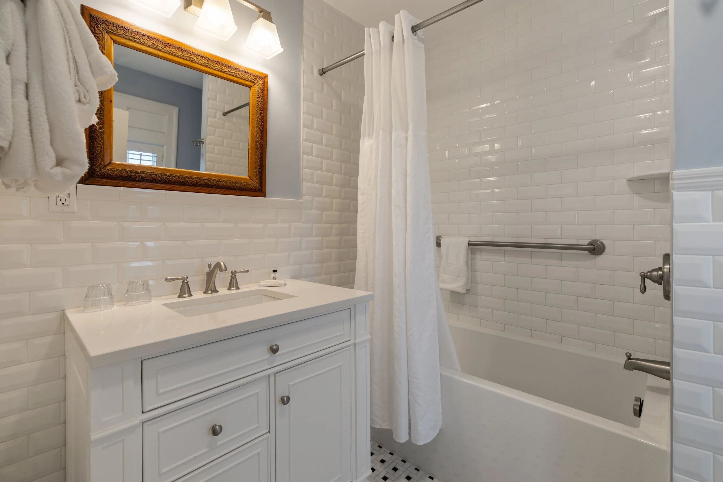 Bathroom with white subway tile walls, a white vanity with a sink, a large mirror with a wooden frame, and a shower area with a white curtain and stainless steel grab bars.