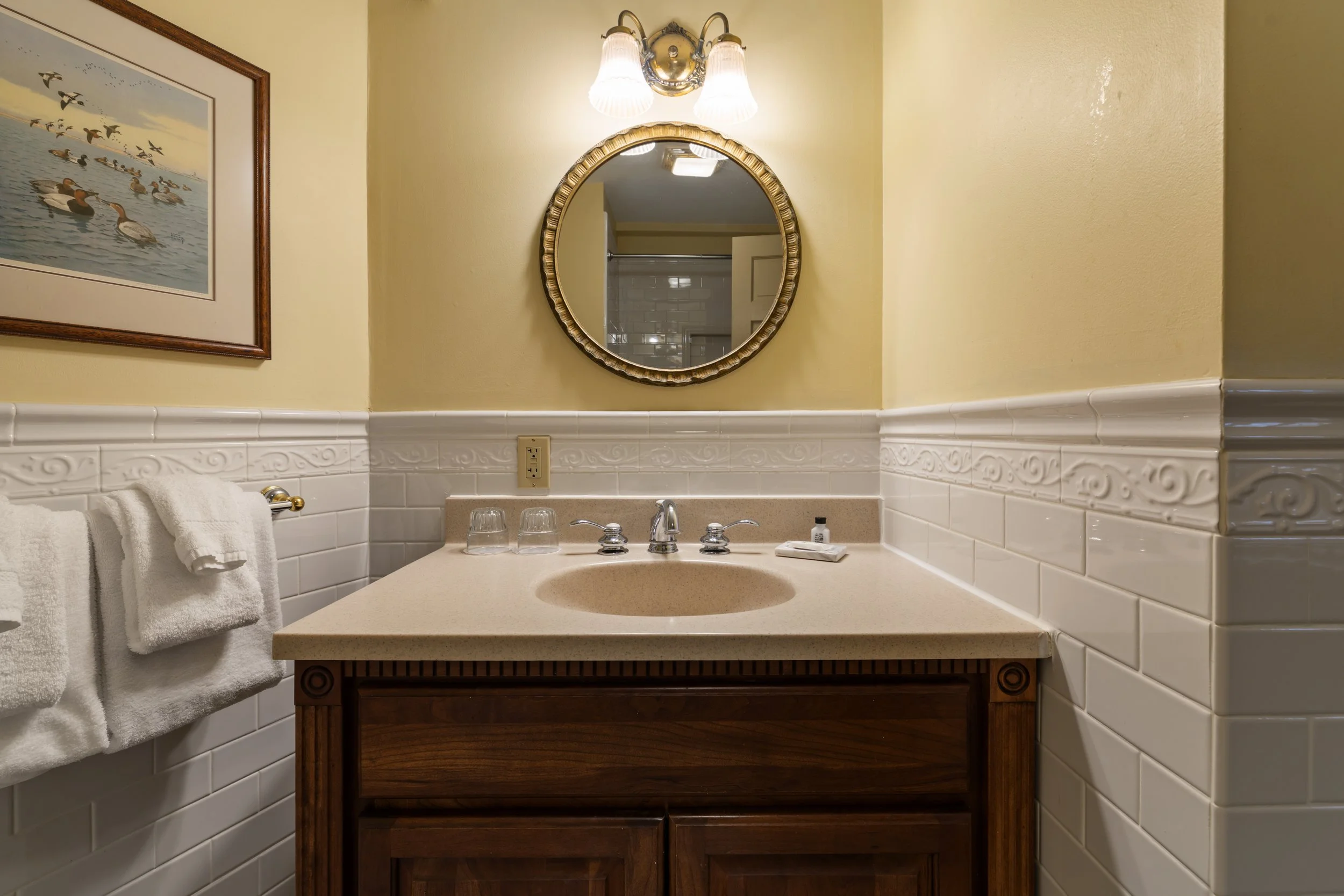 Bathroom vanity area with round mirror, wall-mounted light fixture, yellow painted walls, white tile wainscoting, beige countertop with attached sink, two glasses and soap, wood cabinet below, and framed artwork of ducks on water on the wall.