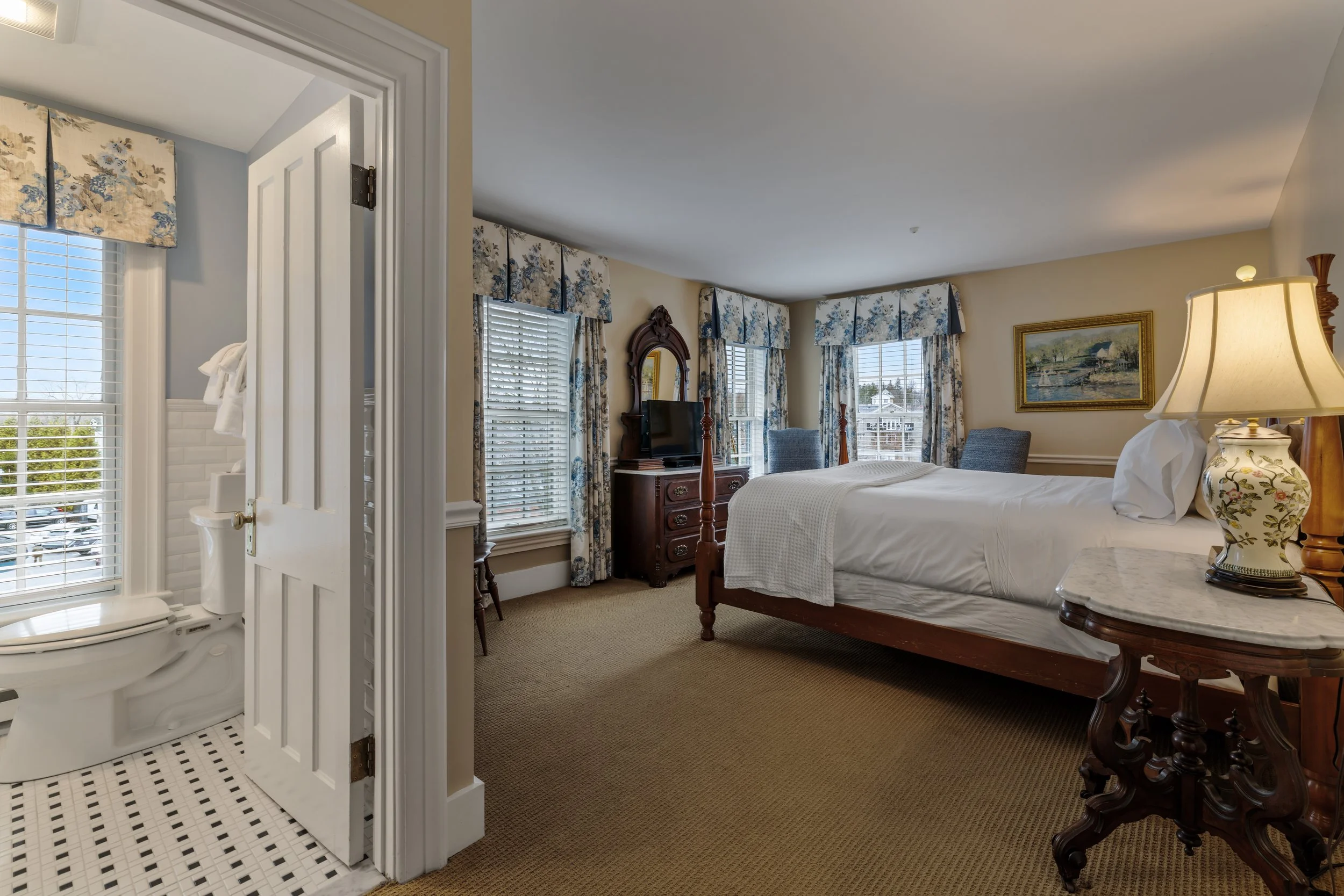 A bedroom with four large windows with floral curtains, a white bed with a white blanket, a wooden dresser with a mirror, a TV, a painting, a lamp, and a small table. A bathroom with a toilet and window is visible through an open door.