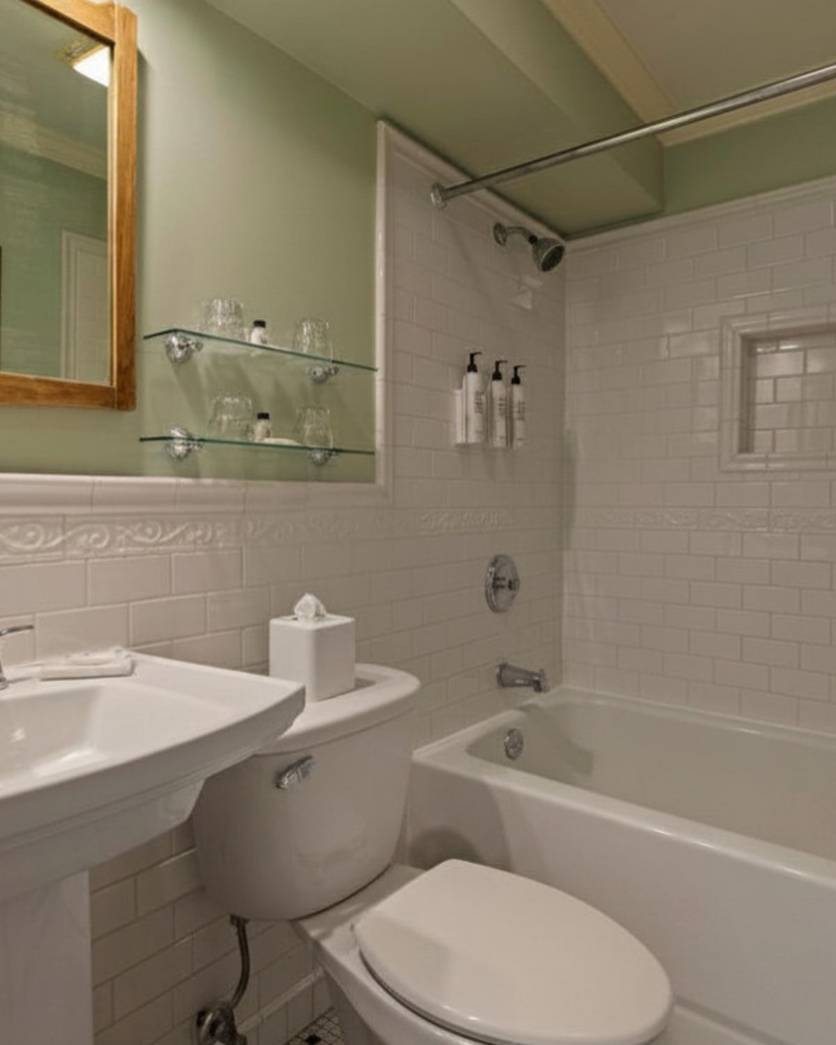 Bathroom with a white bathtub, toilet, and sink. Green wall with mirror and glass shelves holding glasses and toiletries. Tiled shower area with shampoo bottles and a small window.