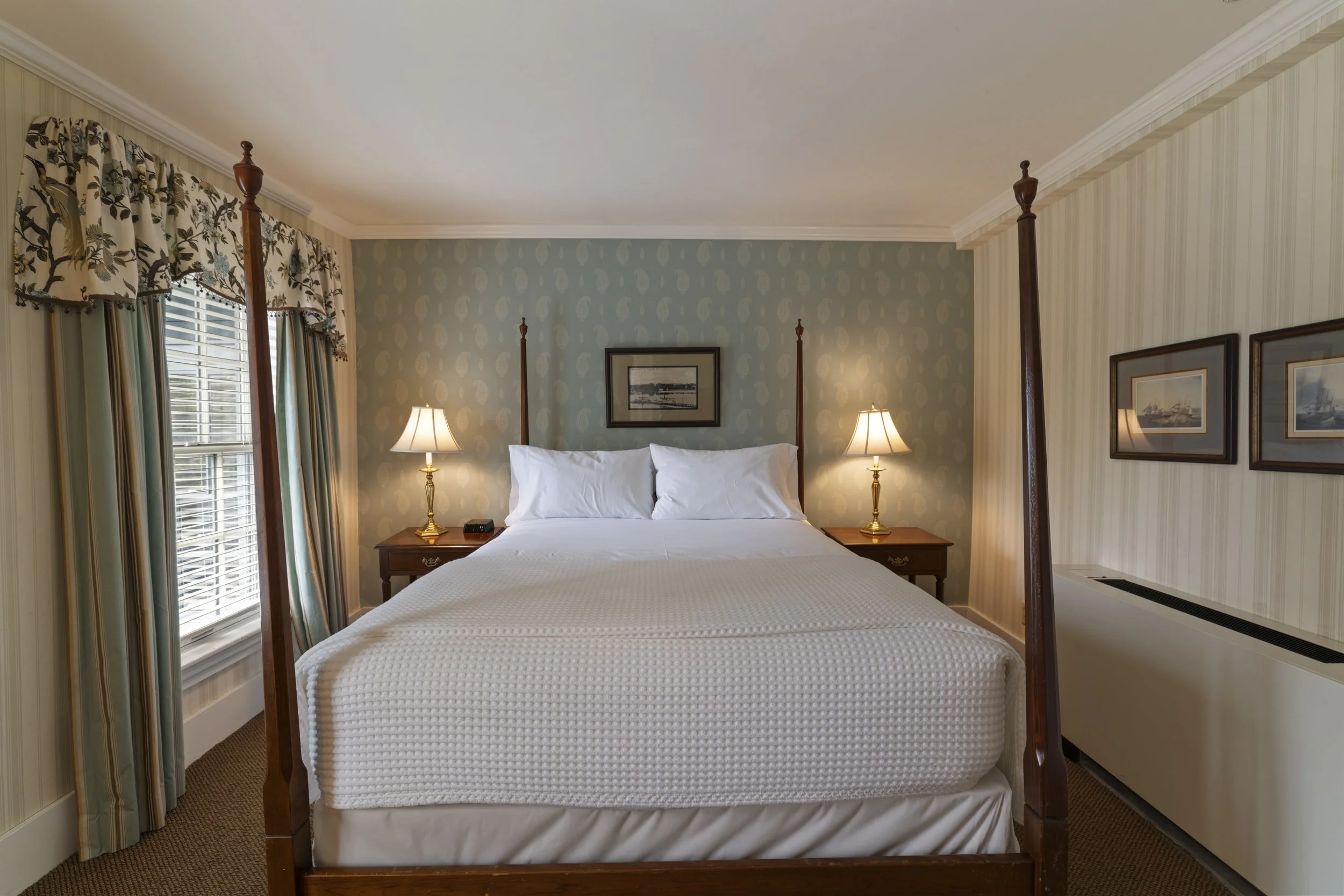 A cozy bedroom with a four-poster bed, white bedding, and two nightstands with lamps. The room has patterned wallpaper, framed artwork, and a window with floral curtains.