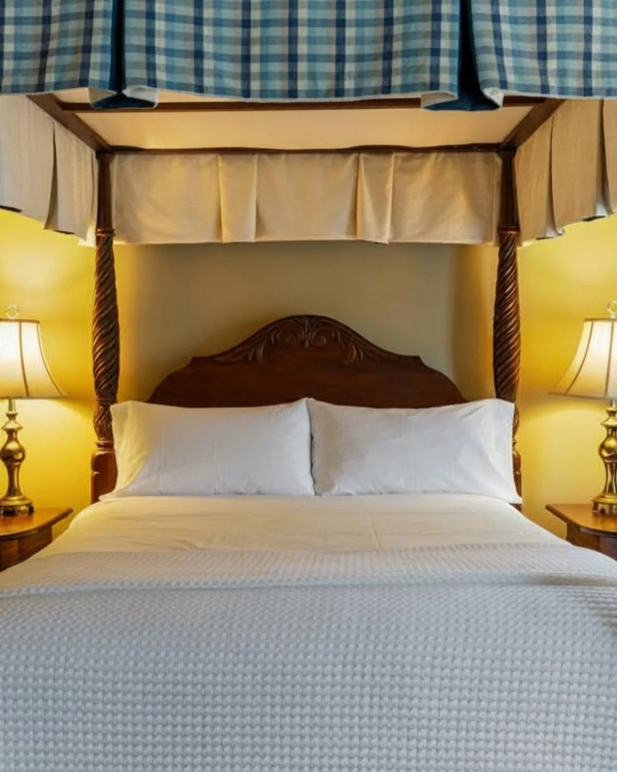 A bedroom with a four-poster bed with a wooden carved headboard, white bedding, and pillows. There are two lamps on bedside tables, and a canopy frame above the bed with blue and cream curtains.