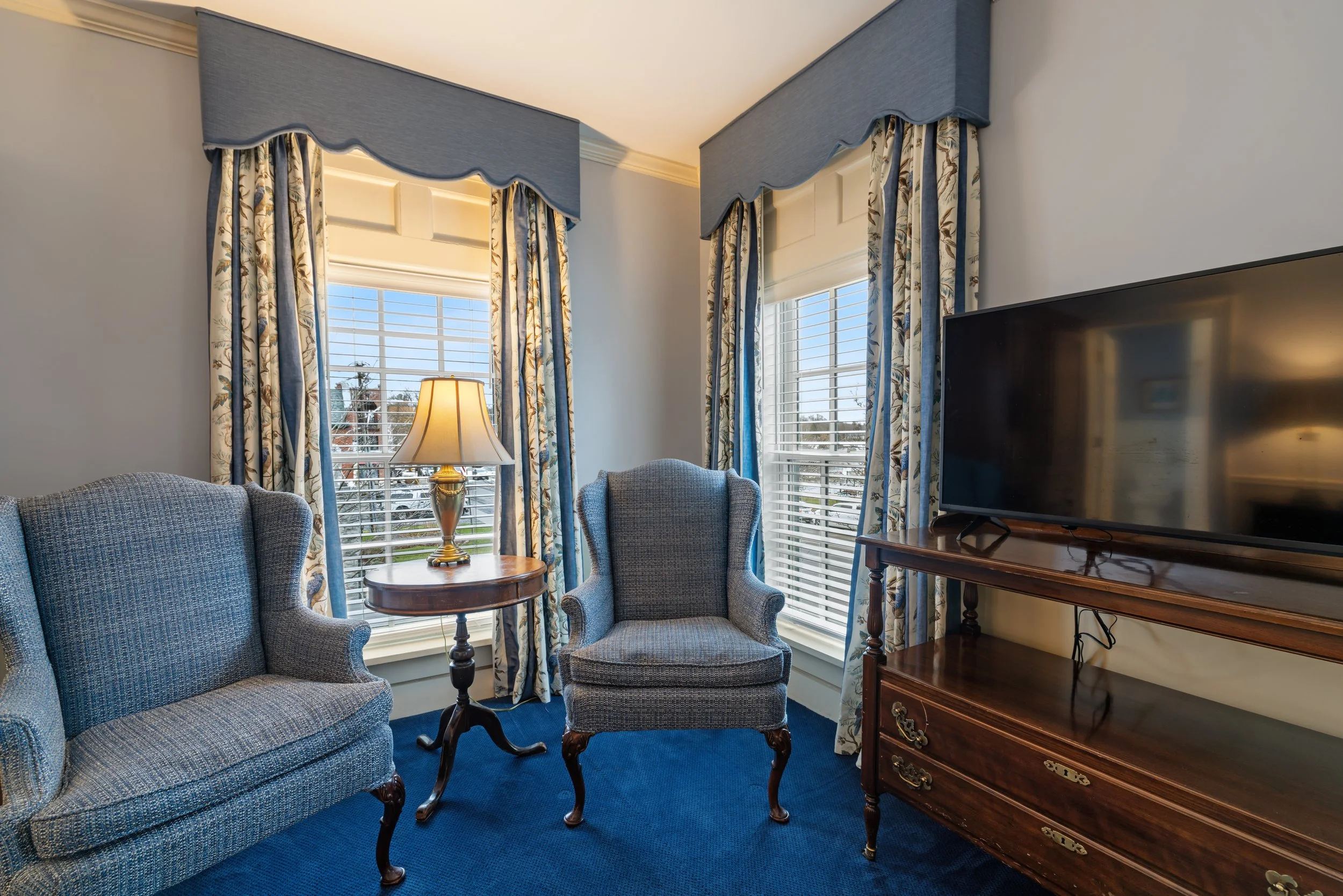 Living room with two armchairs, a small wooden table, a lamp, and a flat-screen TV, all near a large window with floral curtains and valances.