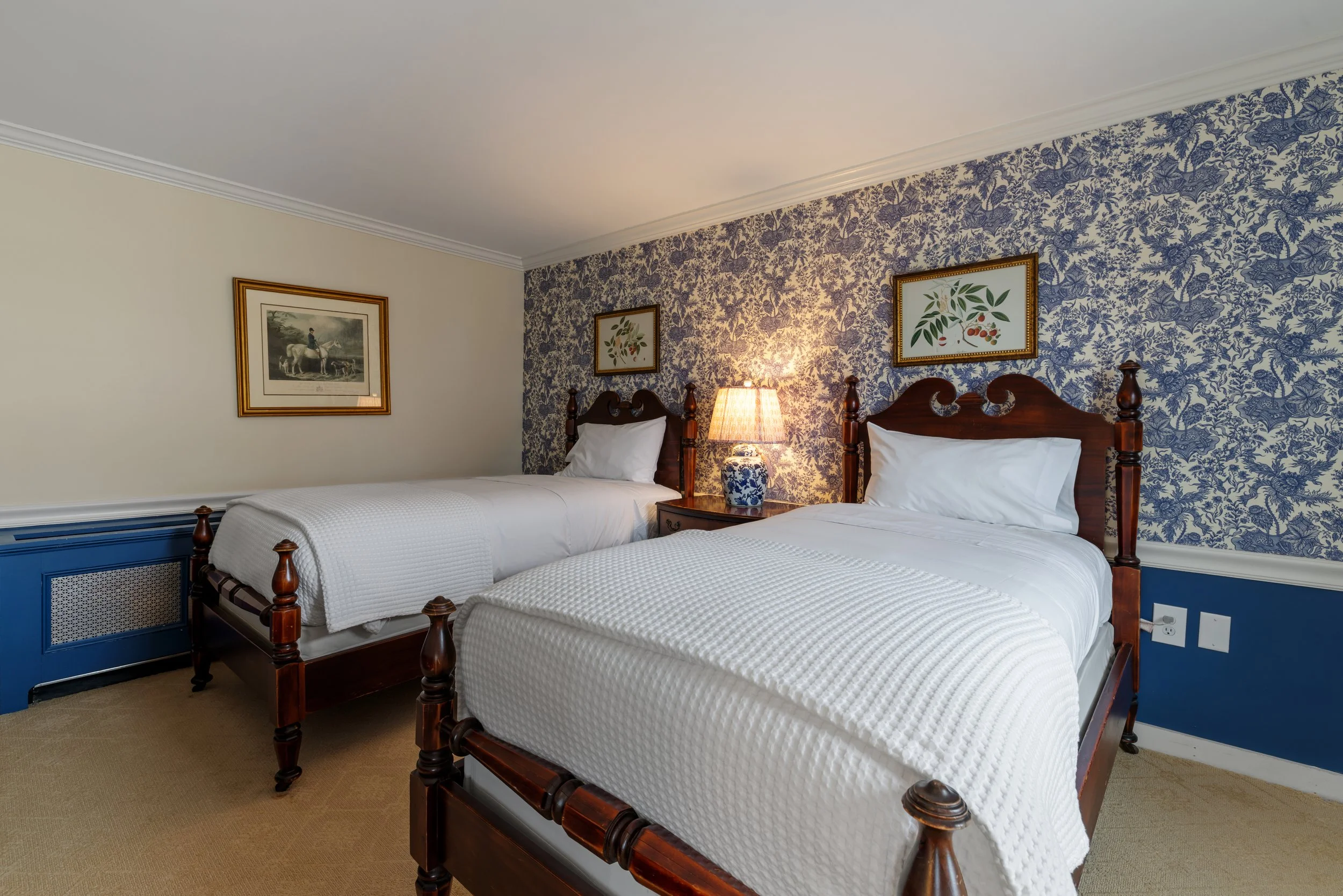 A hotel bedroom with two twin beds, wooden headboards, white bedding, a nightstand with a lamp, framed artwork on the walls, and blue and floral patterned wallpaper.