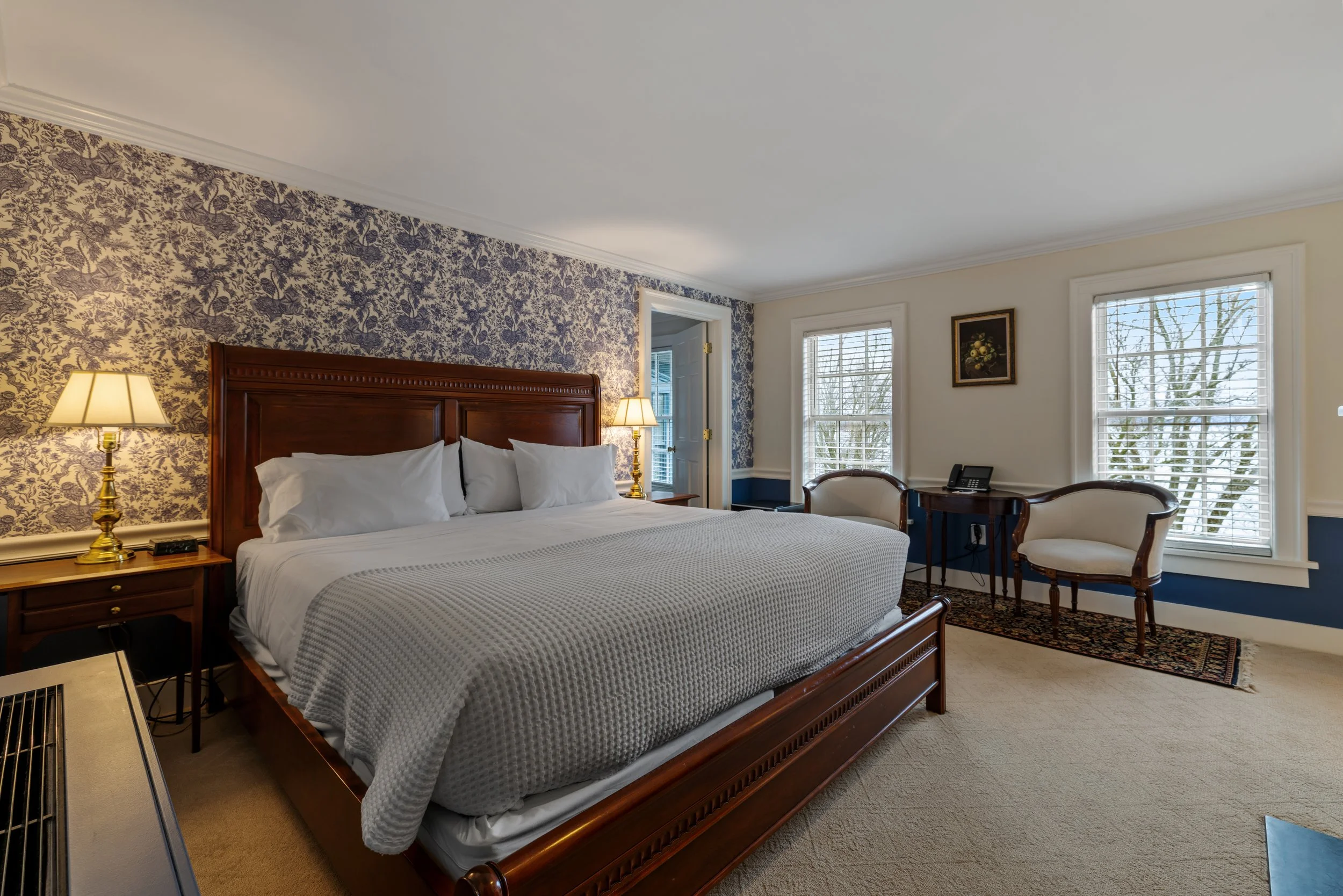 A hotel room with a large wooden bed, white bedding, two lamps on nightstands, windows with blinds showing trees outside, and a sitting area with chairs and a small table.